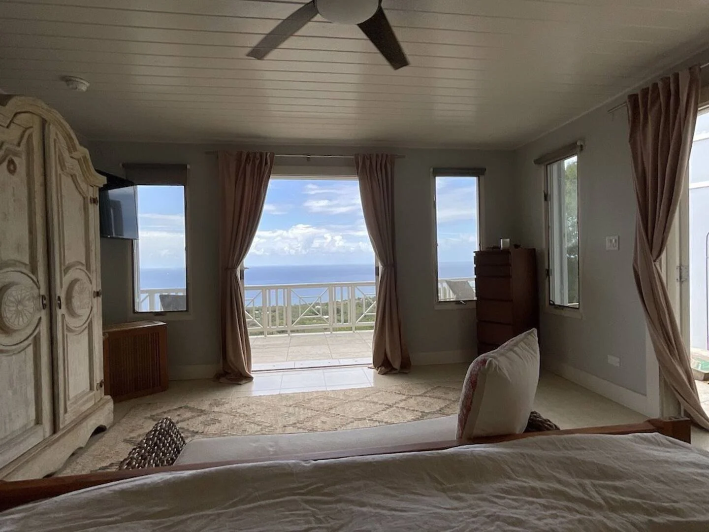 A bedroom with large windows and sliding glass door opening to a balcony, revealing a view of the ocean and blue sky. The room is furnished with an armoire, a dresser, a bed, and a chair, with beige curtains and a ceiling fan.