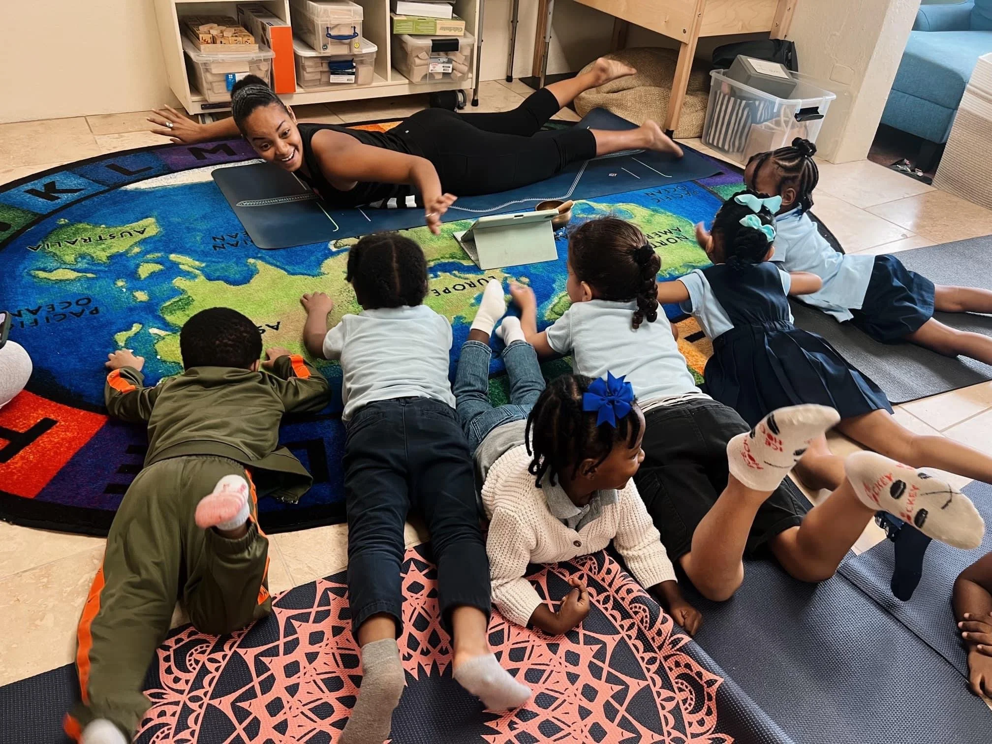 A woman leading a group of young children in a yoga class on a colorful world map rug, with some children lying on their stomachs and one on her side, engaging in a stretch or pose.