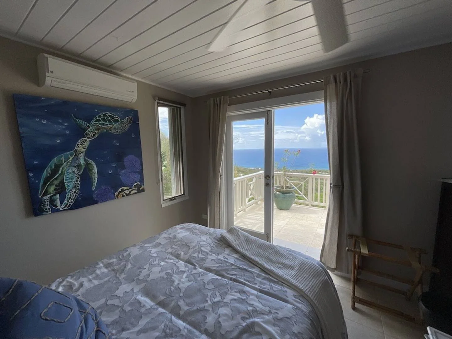 Bedroom with a view of a balcony overlooking the ocean, featuring a bed, a painting of a sea turtle, a window, sliding glass door, potted plant, and beige curtains.