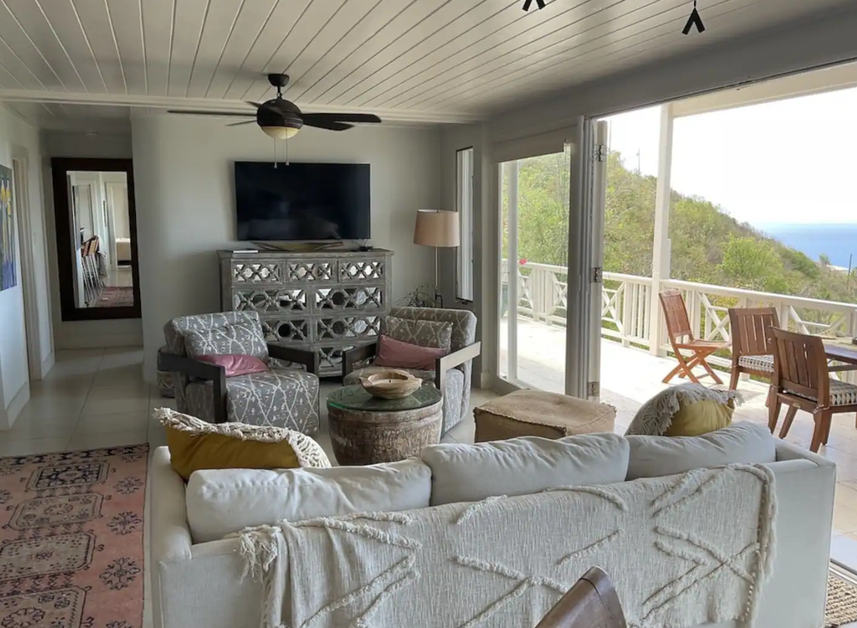 Living room with a white sofa, patterned armchairs, a TV mounted on the wall, and a sliding glass door leading to a balcony with outdoor furniture and a view of trees and the ocean.