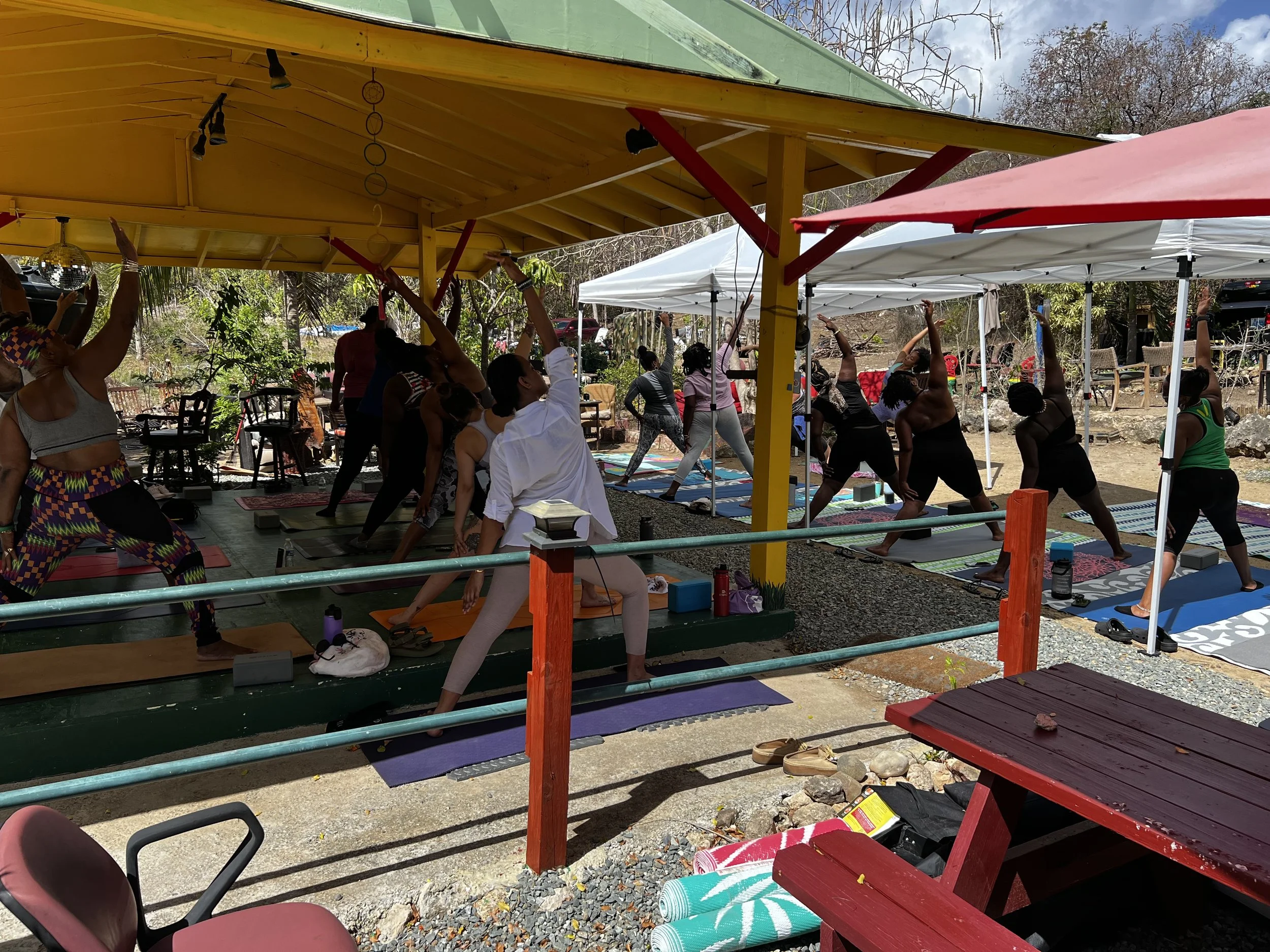 Group of people participating in a daytime outdoor yoga class under a yellow pavilion with trees and outdoor furniture nearby.