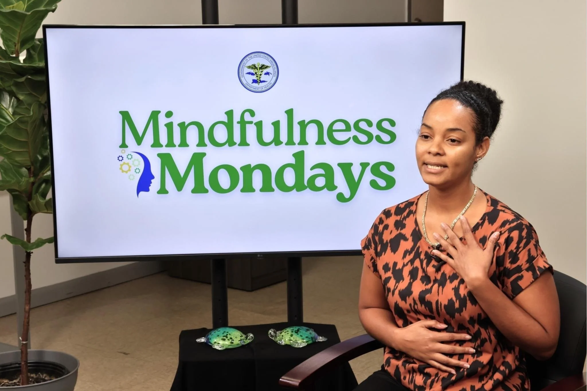 A woman sitting in front of a large screen that reads "Mindfulness Mondays" with a logo at the top. She has her hand on her chest and appears to be speaking.