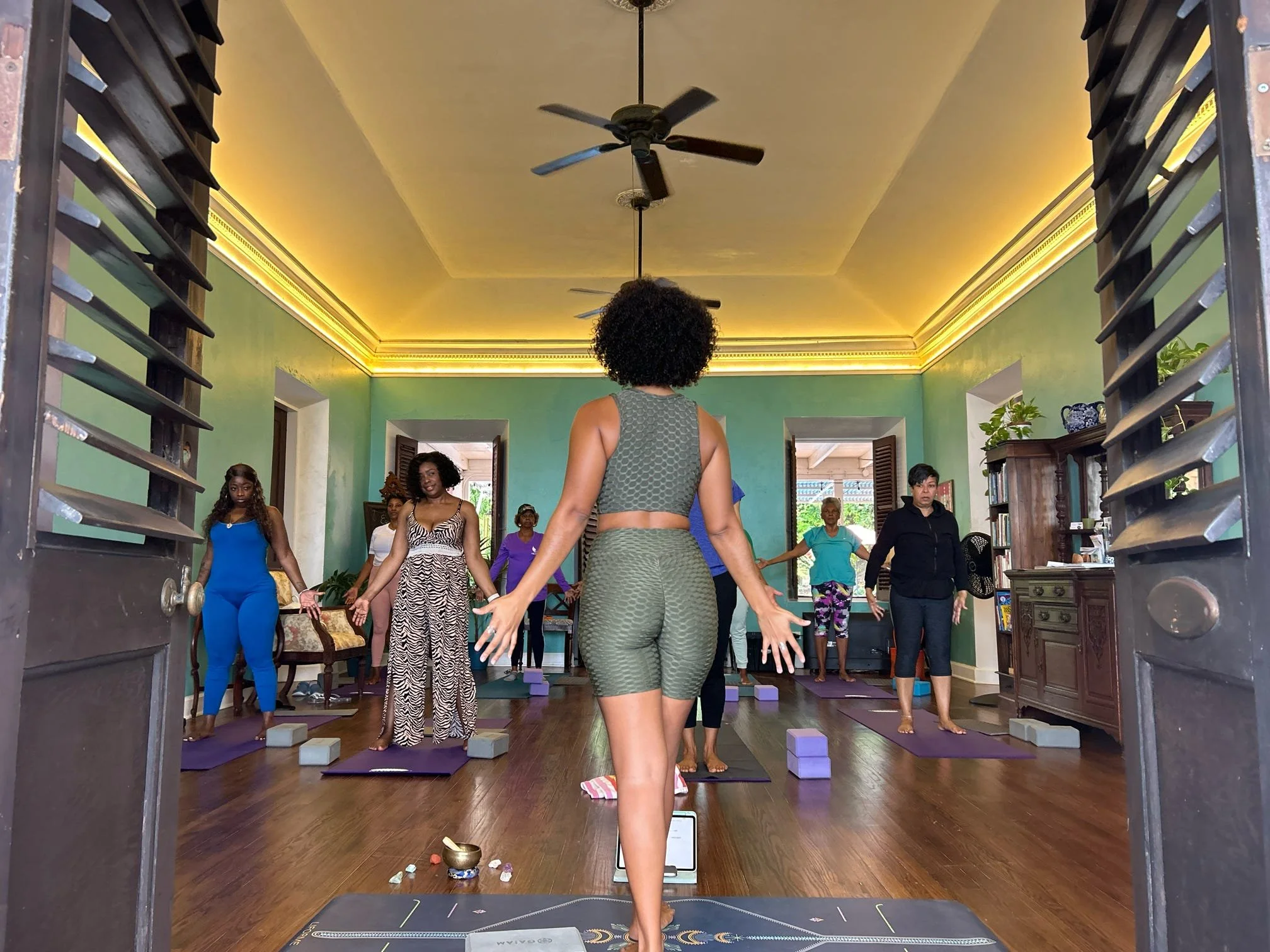 A group of diverse women participating in a yoga class in a bright, spacious room with wooden floors, green walls, and large windows, led by an instructor at the front.