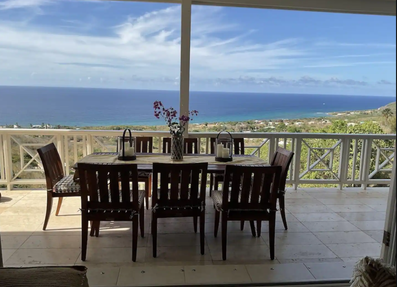 Outdoor dining area with a wooden table and six chairs, overlooking an ocean view with a partly cloudy sky, distant island, and lush green landscape.