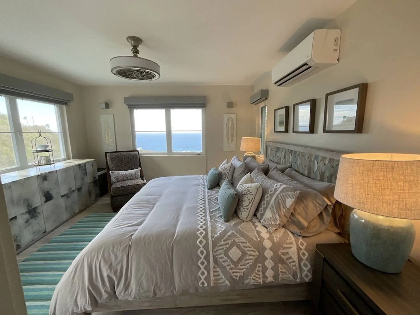 Beachfront bedroom with large bed, multiple pillows, wooden nightstand with lamp, armchair, window view of ocean, coastal decor.