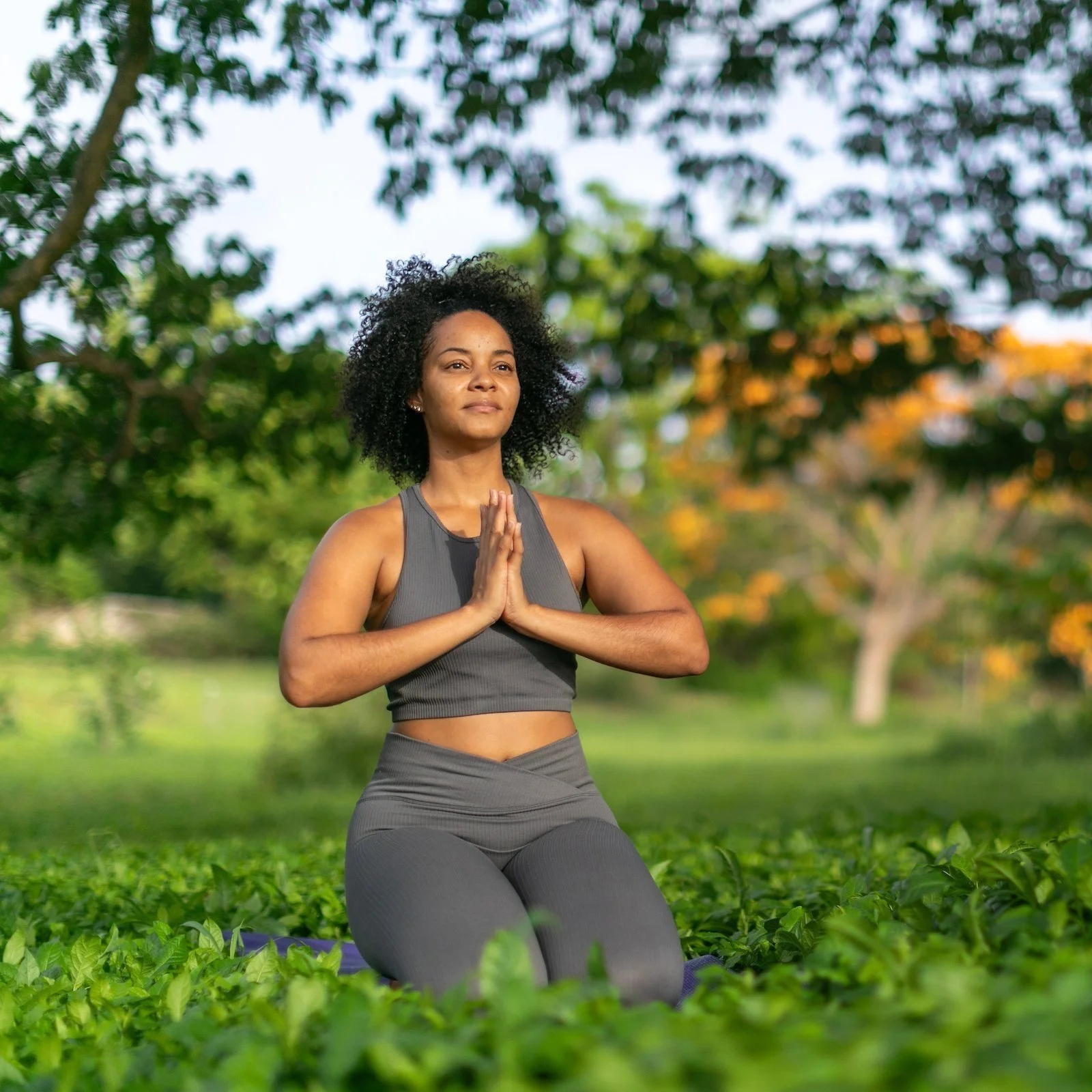 A woman practicing yoga outdoors on her knees with hands in prayer position, surrounded by green grass and trees with orange flowers in the background.