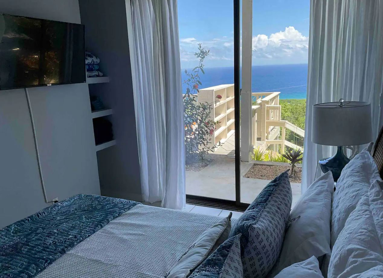 Bedroom with a large sliding glass door opening to an ocean view, a bed with white and patterned pillows, a bedside lamp, and a balcony with stairs leading to greenery and the sea.