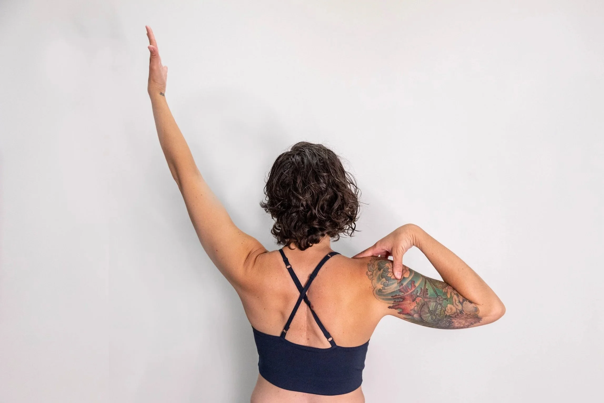 A woman with curly brown hair and tattoos on her arm is standing against a plain white wall, wearing a dark blue strappy top, with one arm raised and the other bent with her hand near her shoulder.