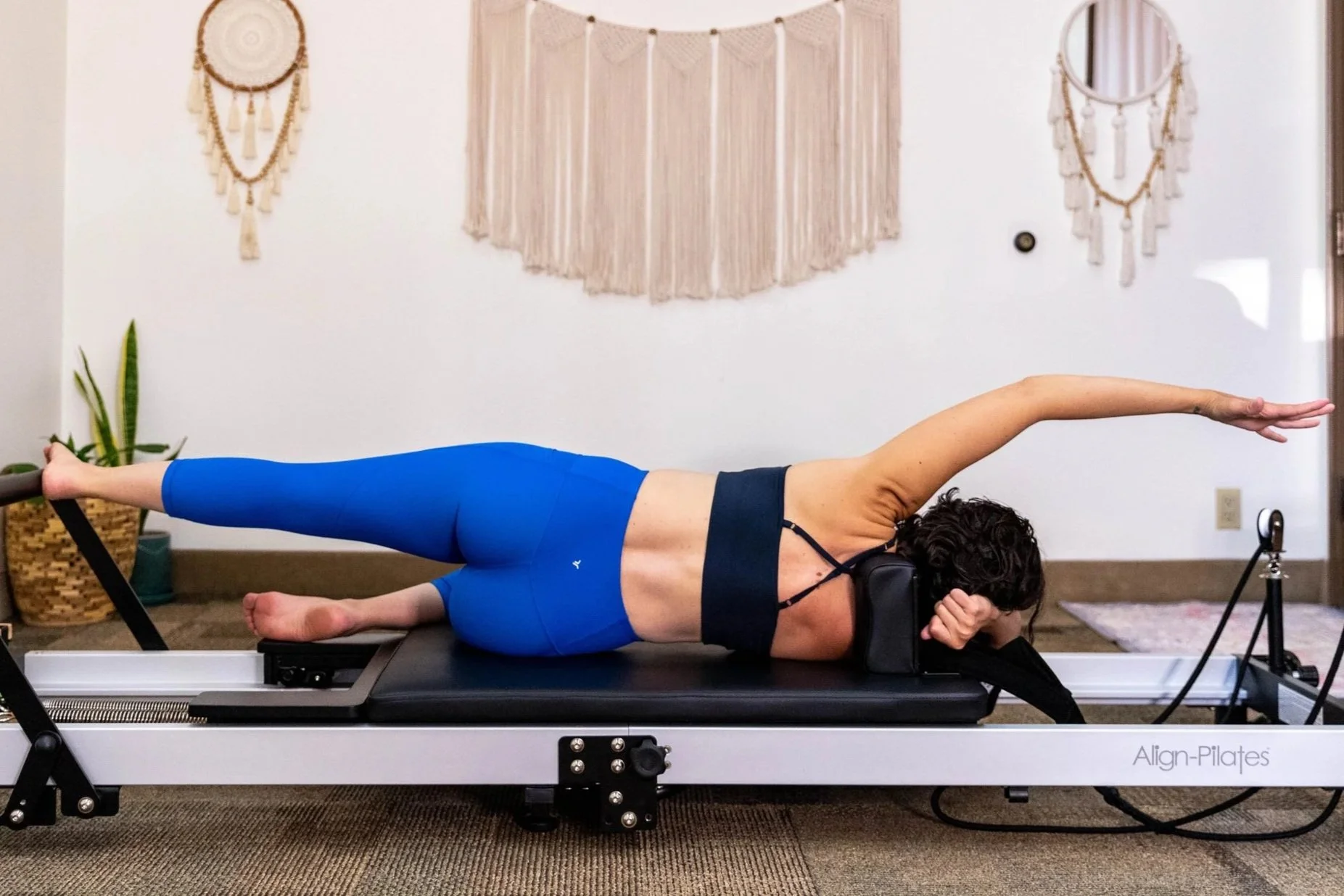 A woman practicing Pilates on an reformer machine, lying on her side with her arm extended upward and her legs straight, in a room decorated with bohemian wall hangings and a potted plant.