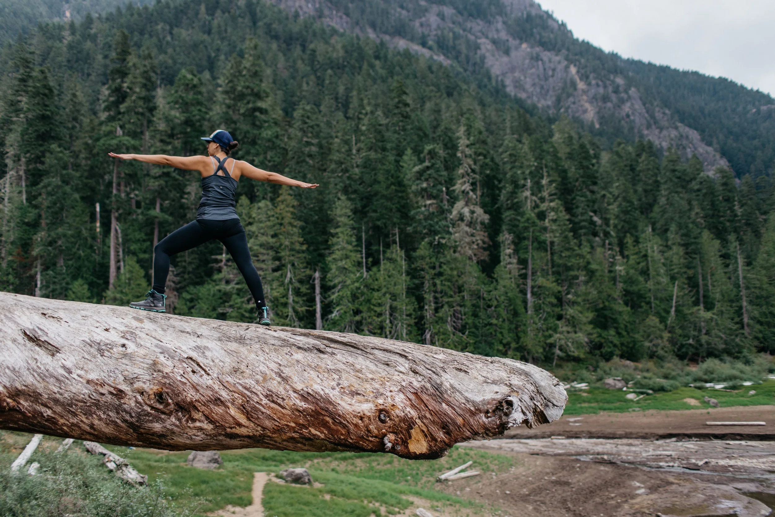A woman in athletic clothing and a baseball cap balancing with arms outstretched on a large fallen tree trunk in a forested mountainous area.