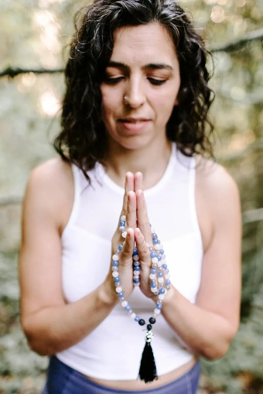 A young woman with curly dark hair and a nose piercing stands outdoors with eyes closed, holding prayer beads in prayer position.