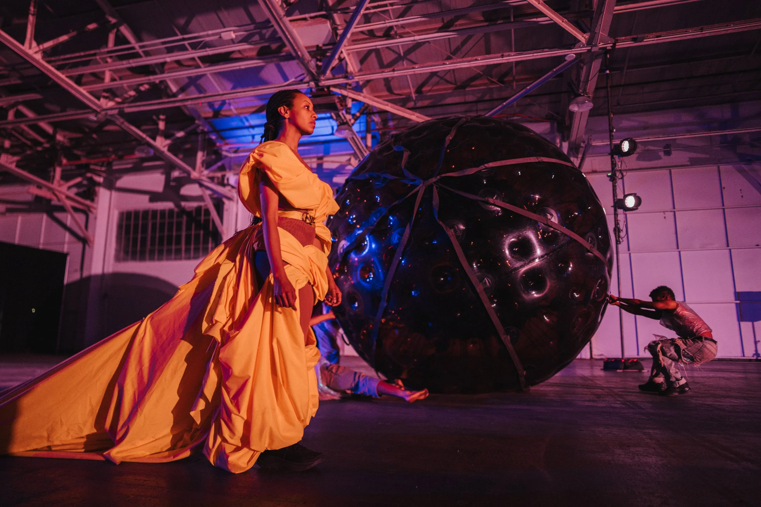 A woman in a flowing yellow dress walking past a large reflective spherical sculpture in an indoor space with industrial ceiling. The scene is illuminated with colorful pink and blue lighting.