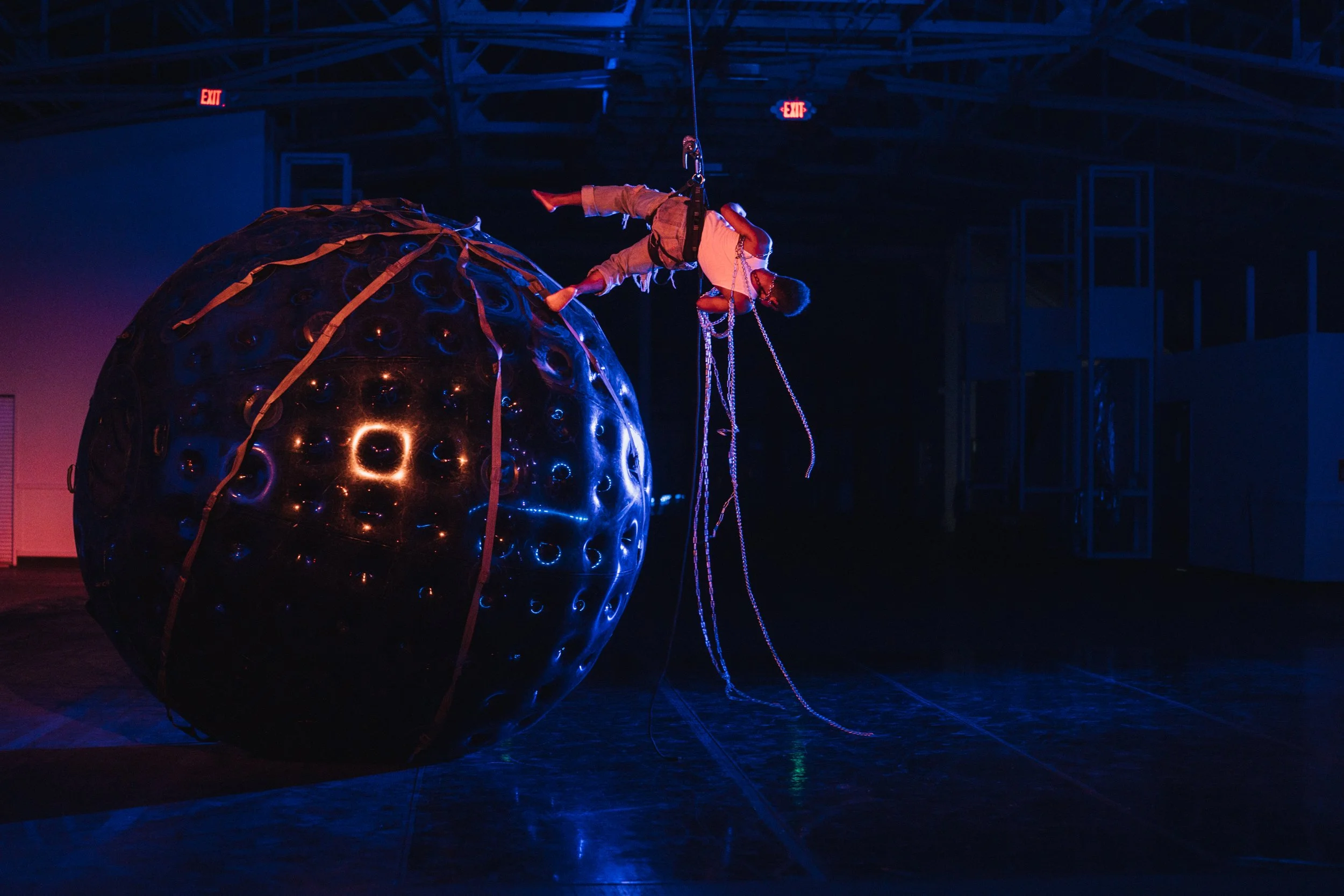 A performance art installation showcasing a person hanging upside down from a chain, above a large, glossy, black spherical sculpture with circular patterns, illuminated by vibrant blue and purple lights in an industrial space with visible structural elements and exit signs.
