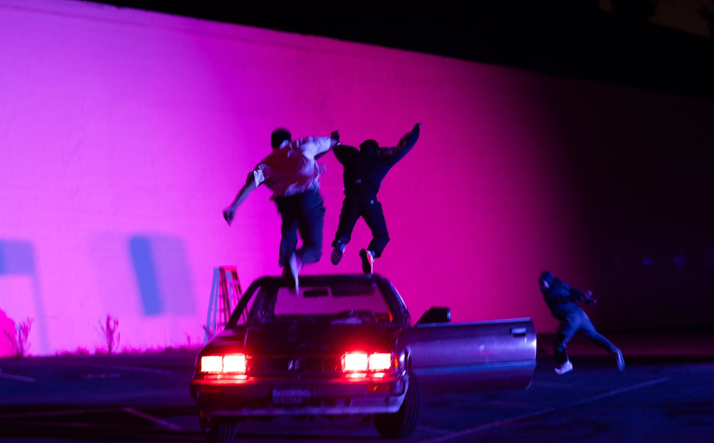 Two people standing on the back of a pickup truck during a neon-lit night, with a pink and purple background, some small trees visible in the distance.