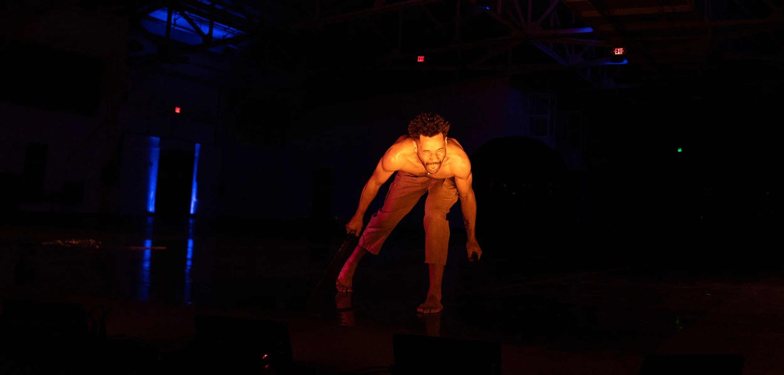A shirtless man with curly hair is in a dance or performance pose on a dark stage with dim lighting and blue and red spotlights.