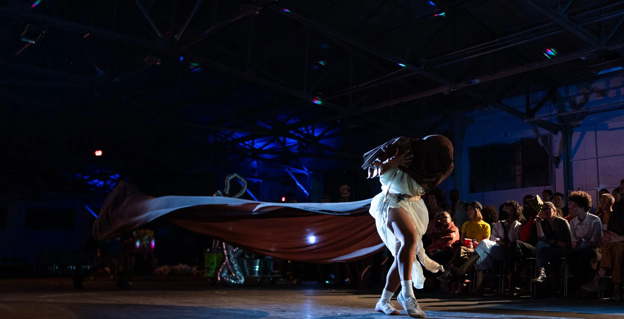 Person performing a dance in a dark indoor venue with audience watching, illuminated by blue lighting. The dancer is dressed in a white costume with a flowing train.
