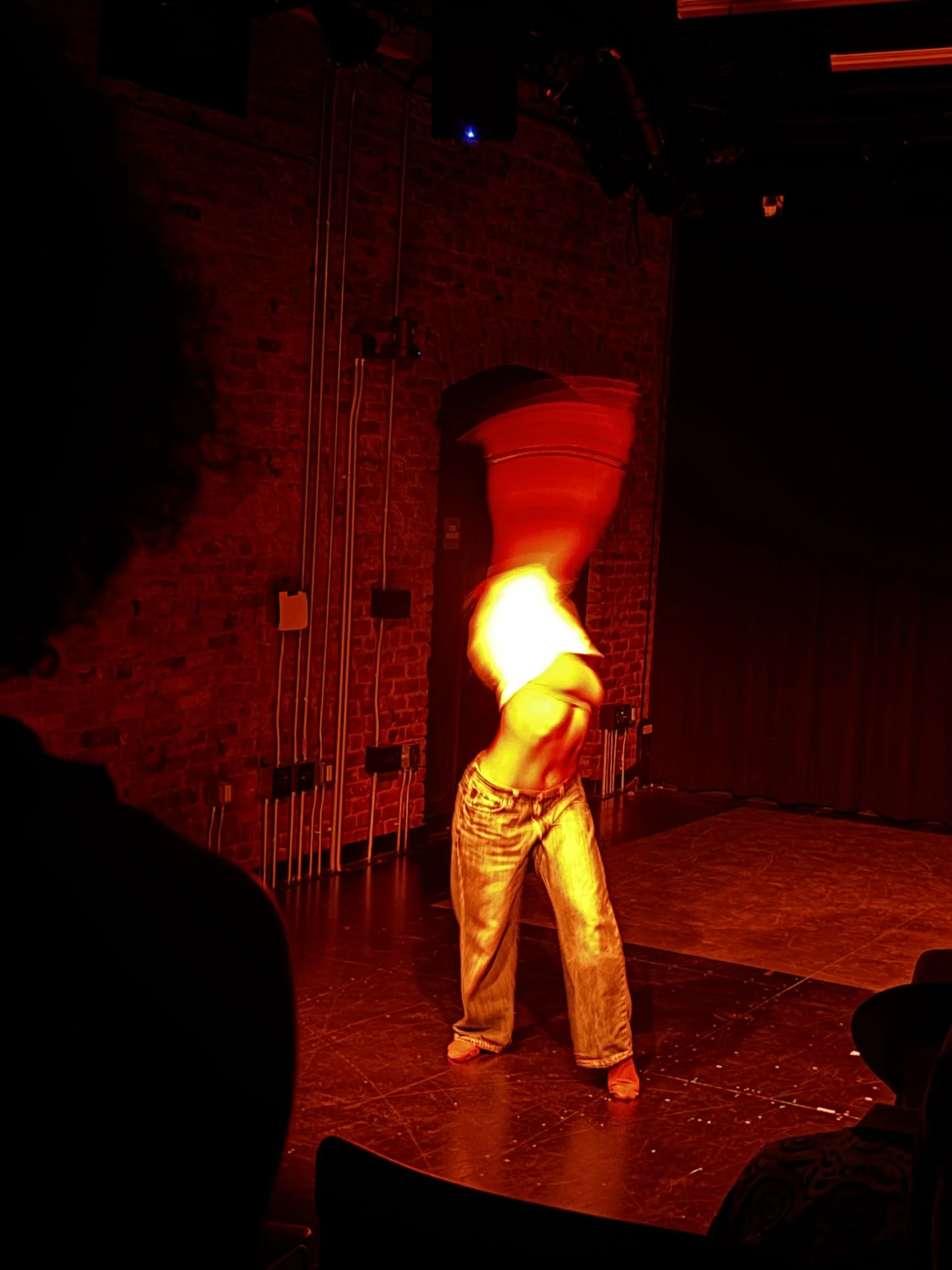 A performer on stage with a fiery prop, wearing casual jeans and a shirt, performing in a dimly lit venue with a brick wall background and stage lighting.