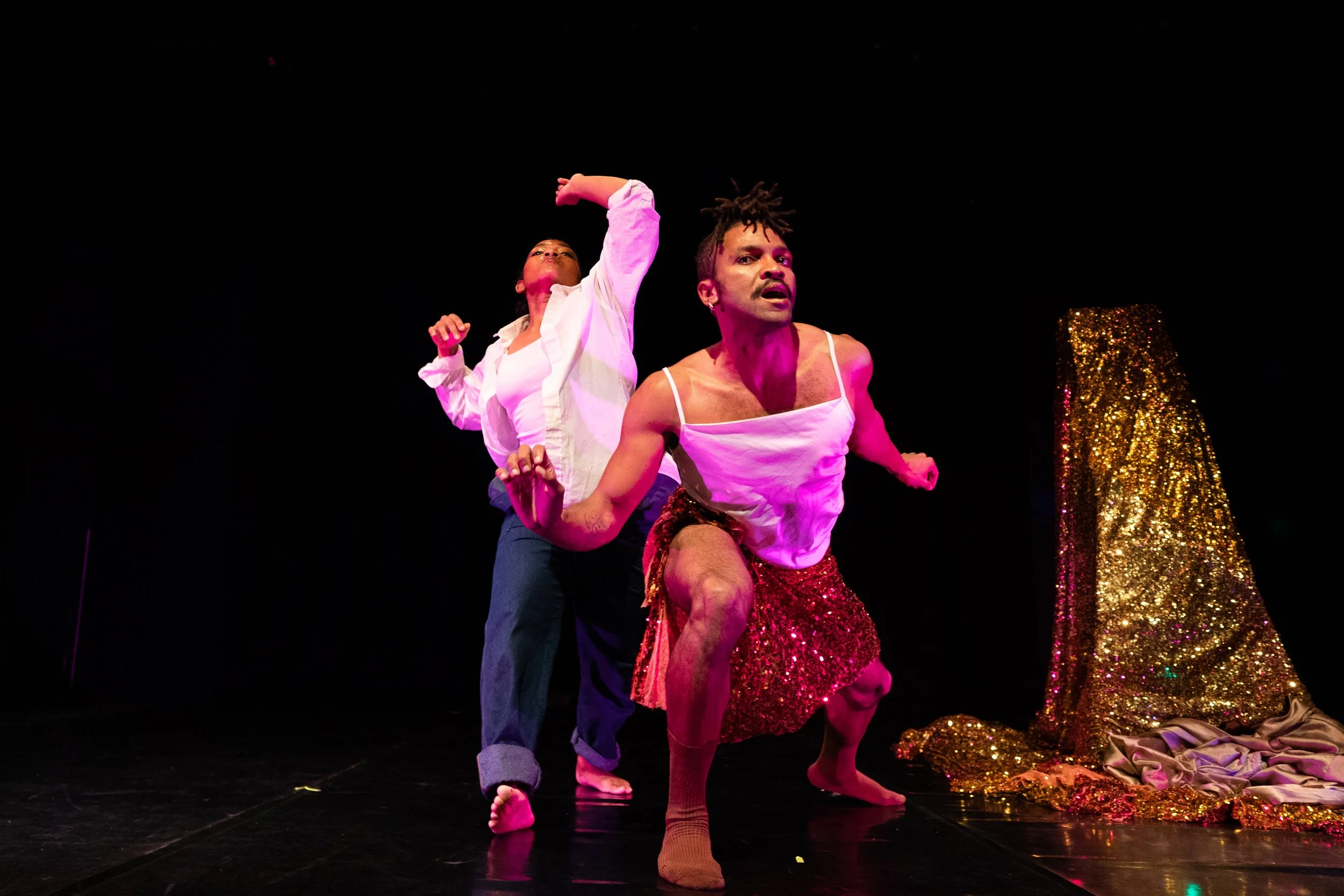 Two performers on stage, one with a raised arm and the other crouching, in front of a gold, glittery backdrop.