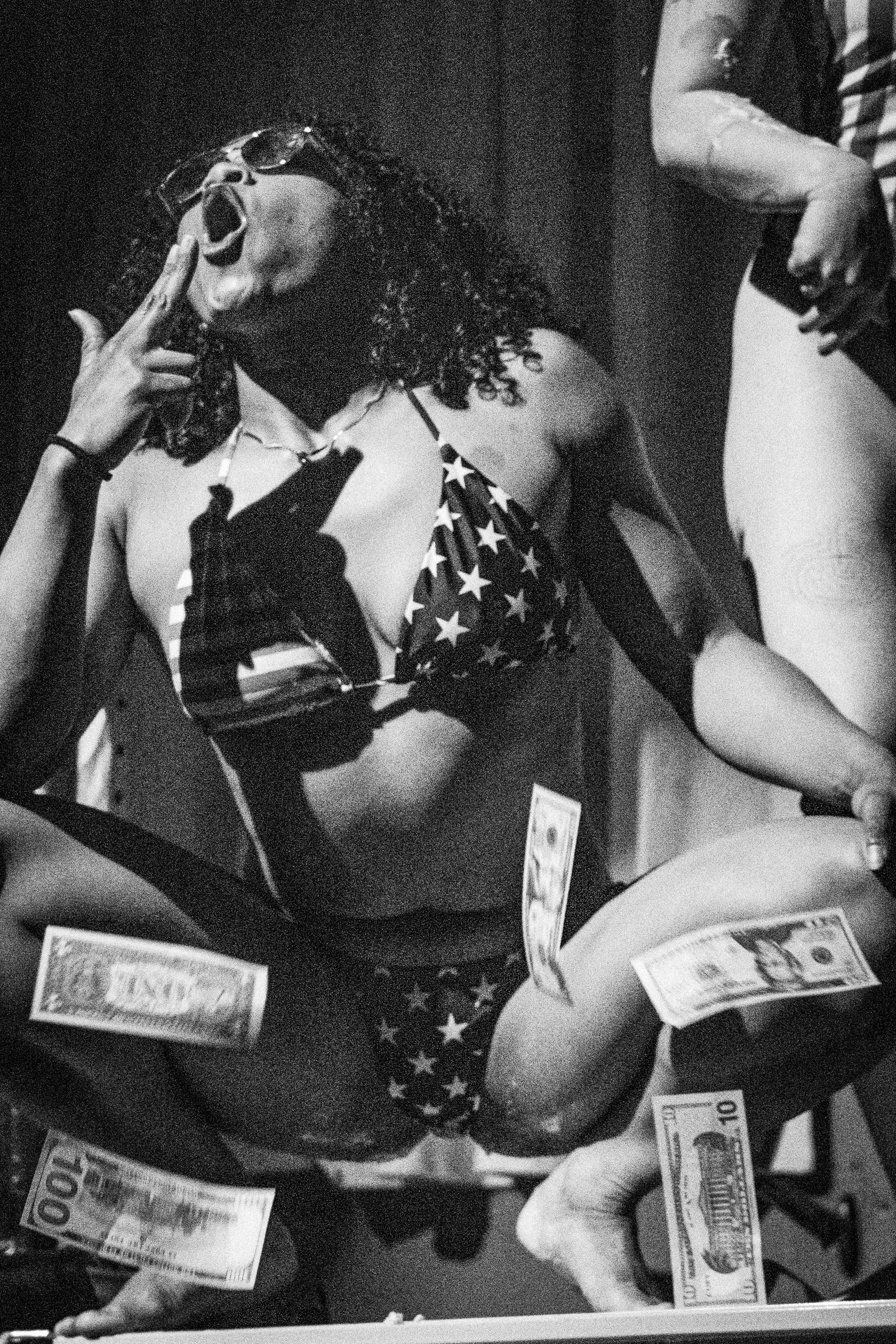 People surrounding a table, counting and handling cash, including one person holding a one hundred dollar bill. There are various bills on the table and some in their hands. The image is in black and white.