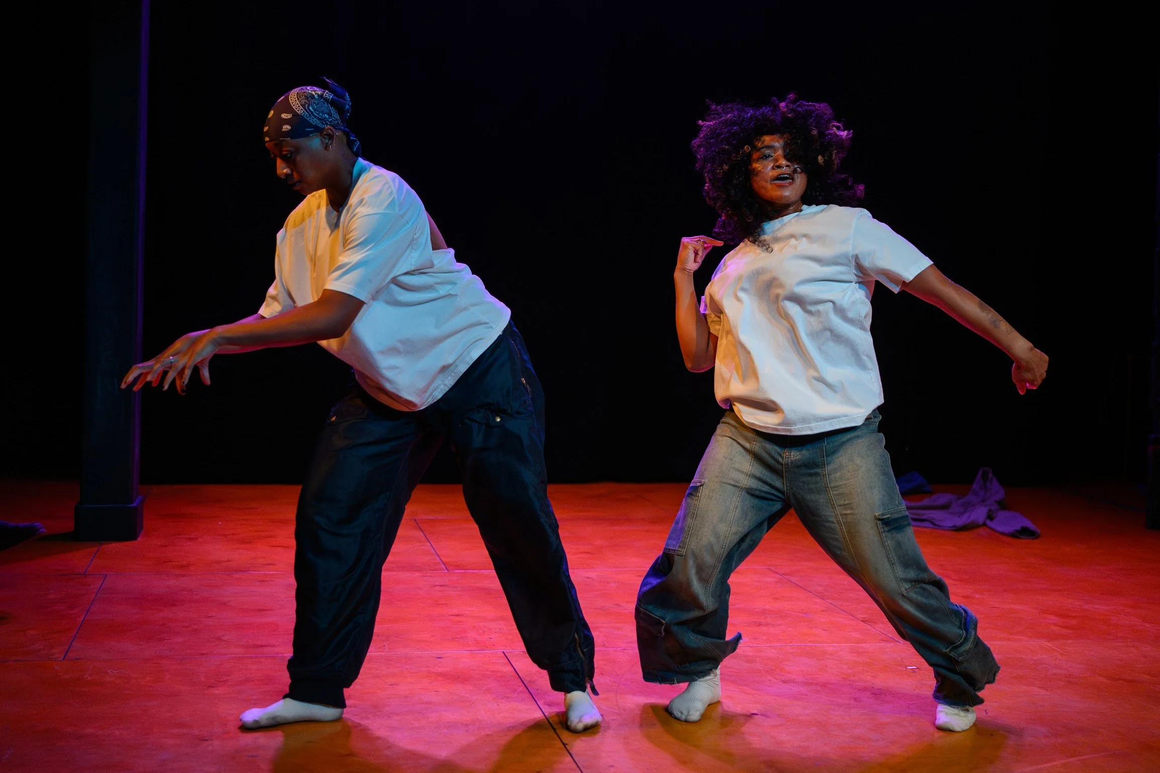 Two women are performing a dance on a stage with a wooden floor and a dark background. They are dressed in casual clothing, including white t-shirts and jeans, and are wearing socks instead of shoes. One woman has a headscarf, and the other has voluminous curly hair. They are in expressive dance poses, with one woman pointing and the other leaning forward.