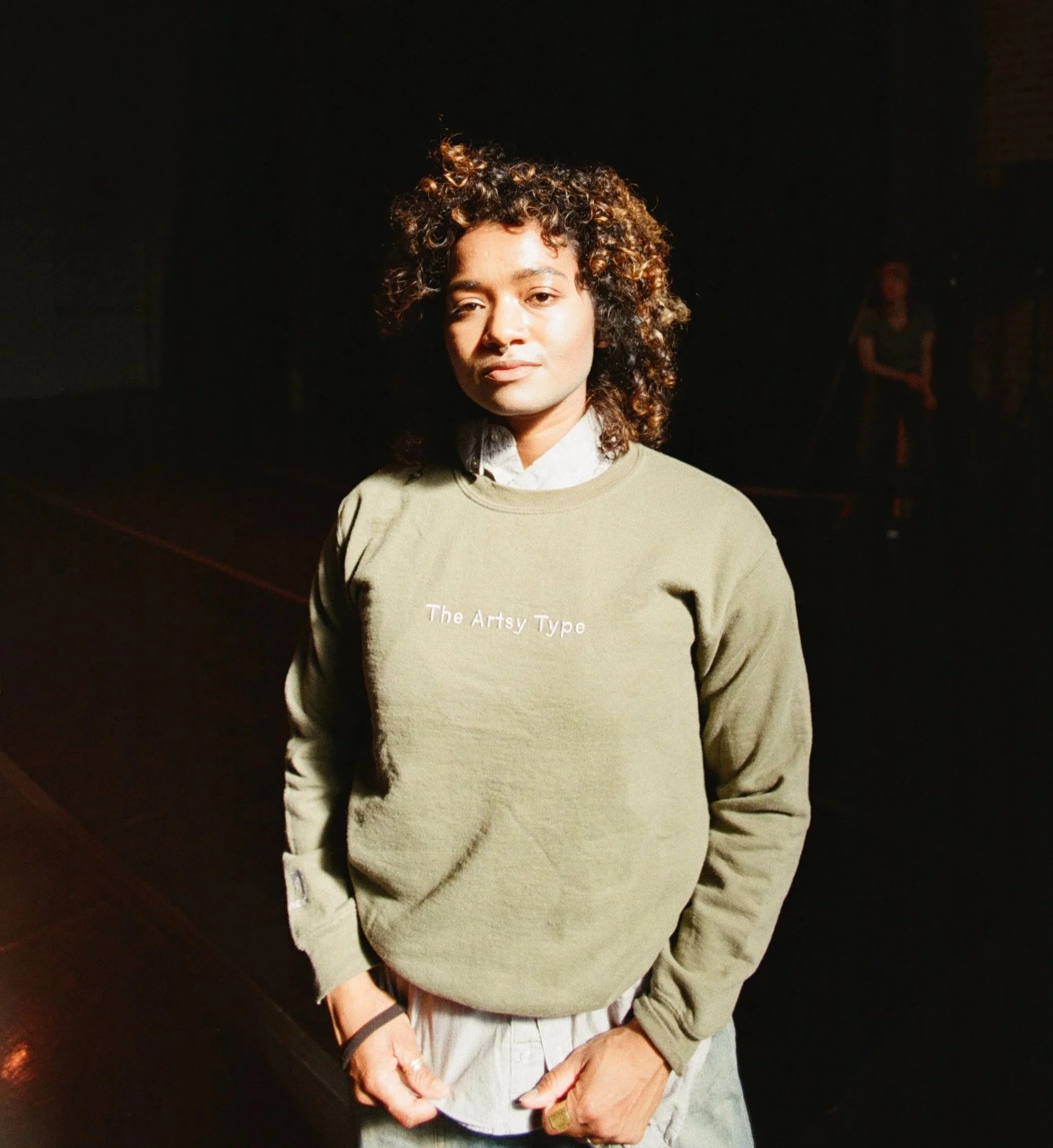 Close-up of a woman with curly hair wearing a beige sweatshirt and light-colored pants, standing in a dimly lit environment.