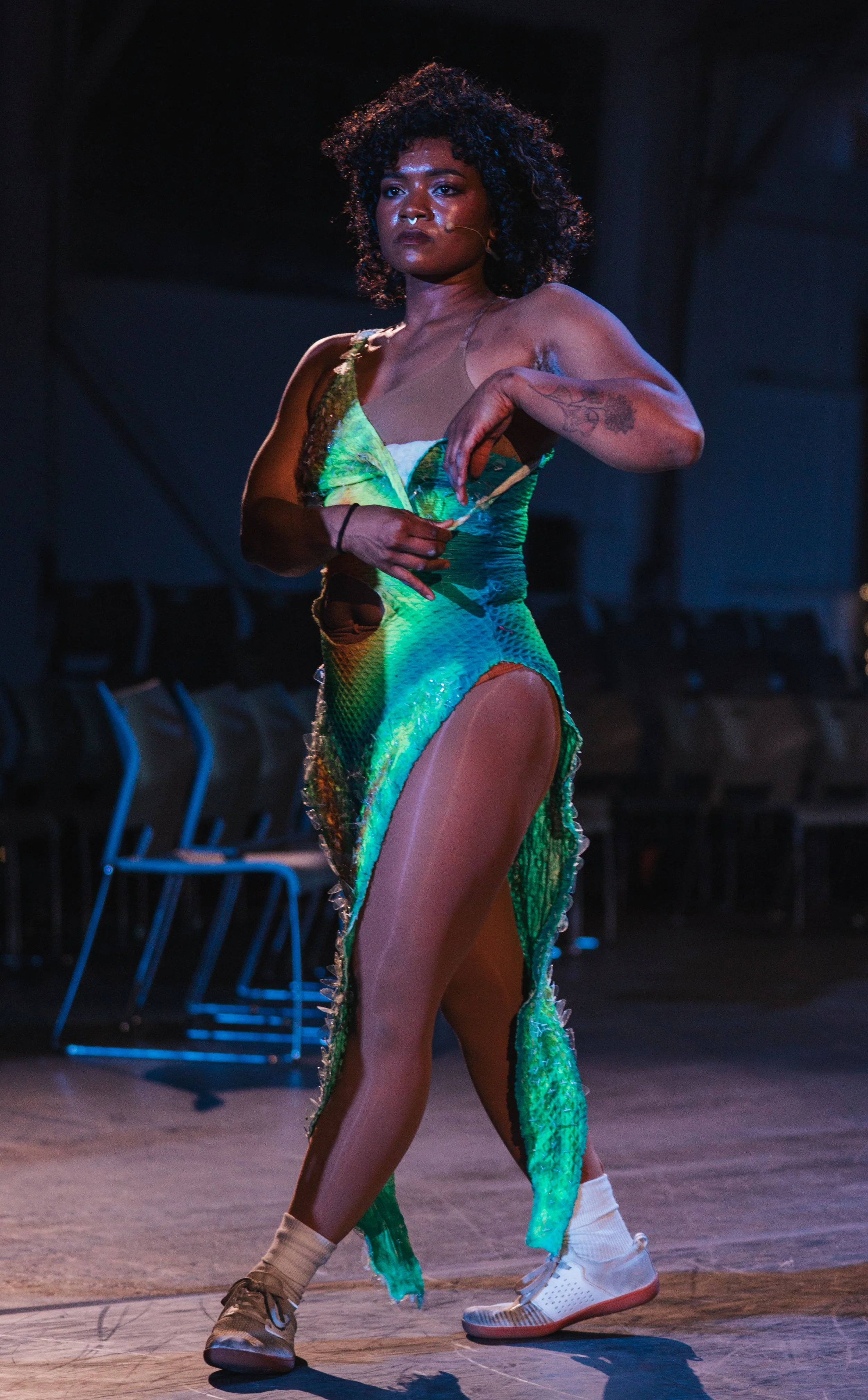 A woman with curly hair in colorful clothing and sneakers performing a dance move on stage.