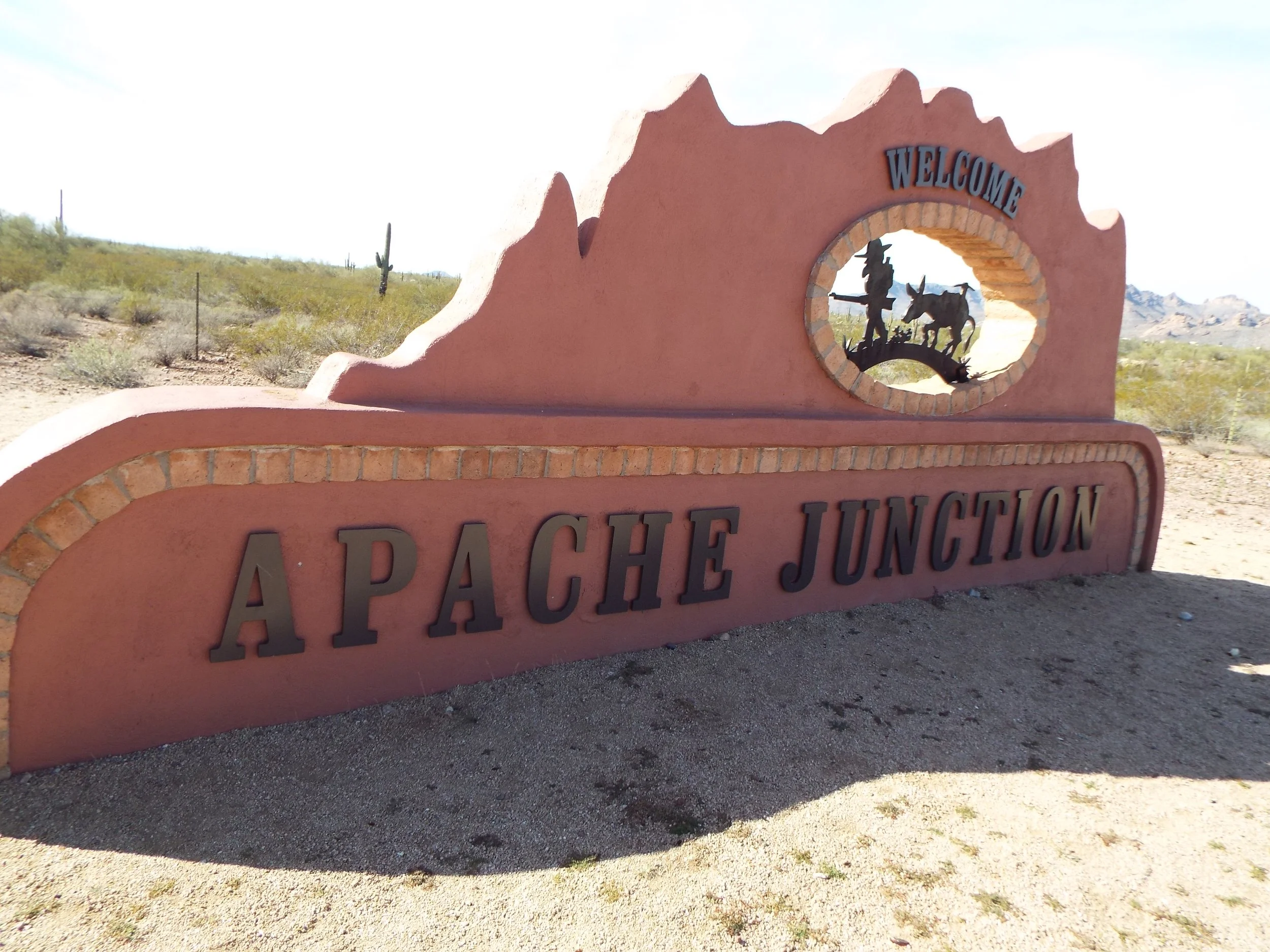 Sign that says 'Apache Junction' with a welcome message and a decorative cutout of a cowboy and a donkey in a desert landscape.