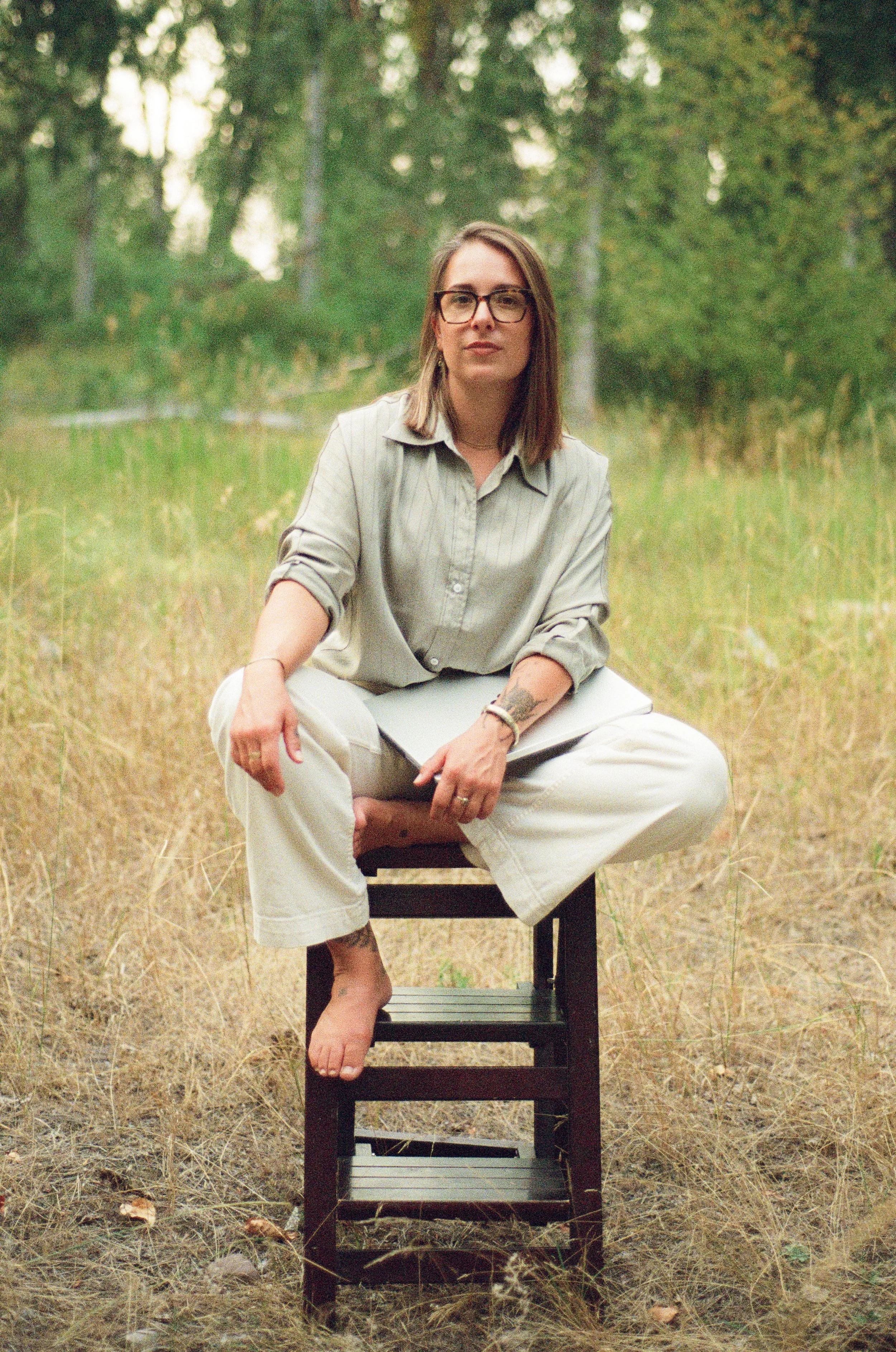 The founder with glasses, wearing a light green shirt and white pants, sitting cross-legged on a chair outdoors in a grassy field with trees in the background. She is holding a notebook on her lap and looking towards the camera.