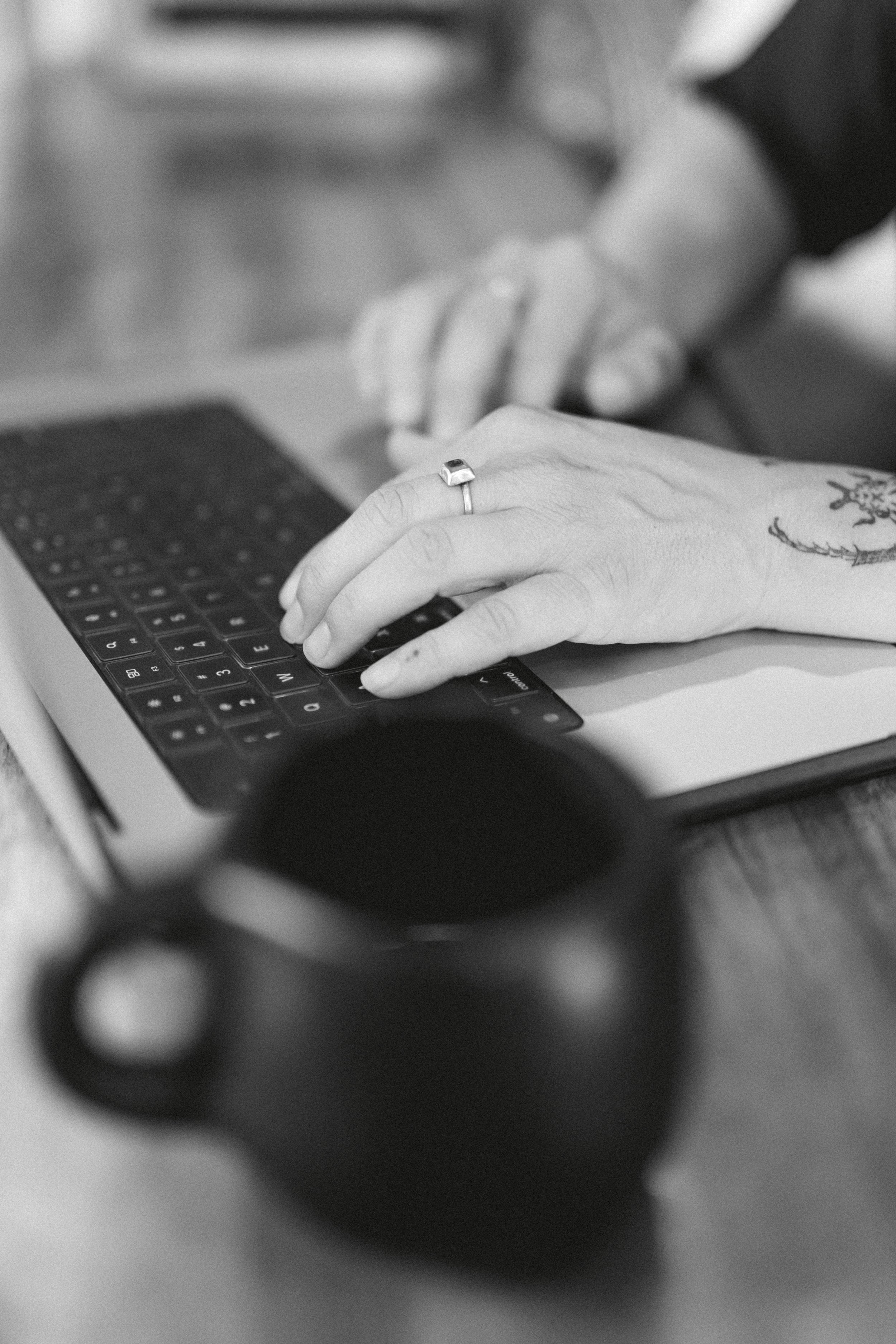 Close-up of a person's hand on a laptop keyboard, with tattoos visible on the wrist, blurred person's face in the background, black and white tone.