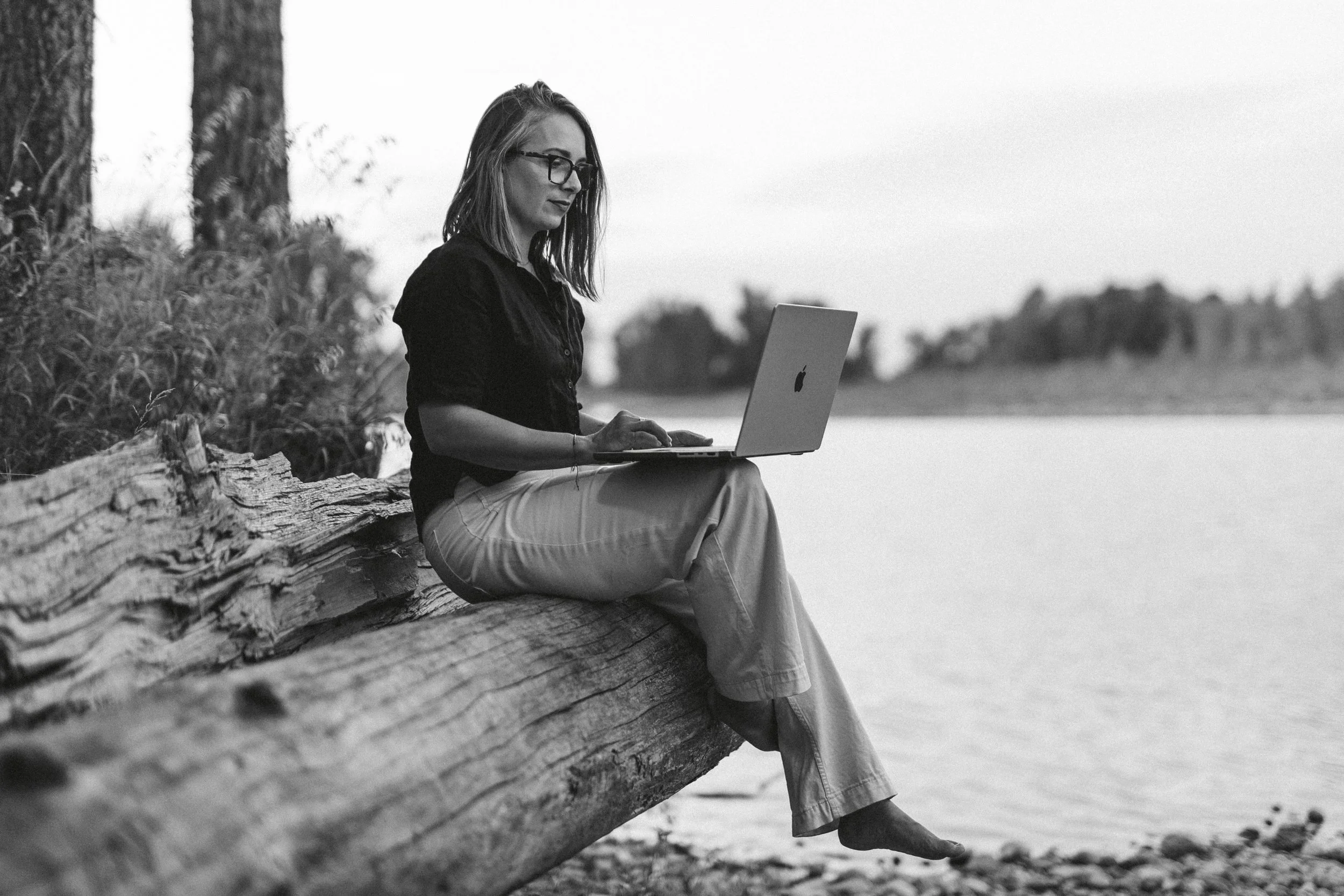 The founder of Studio Aissa with glasses sitting on a log by a body of water, using a laptop, in a black and white outdoor setting.