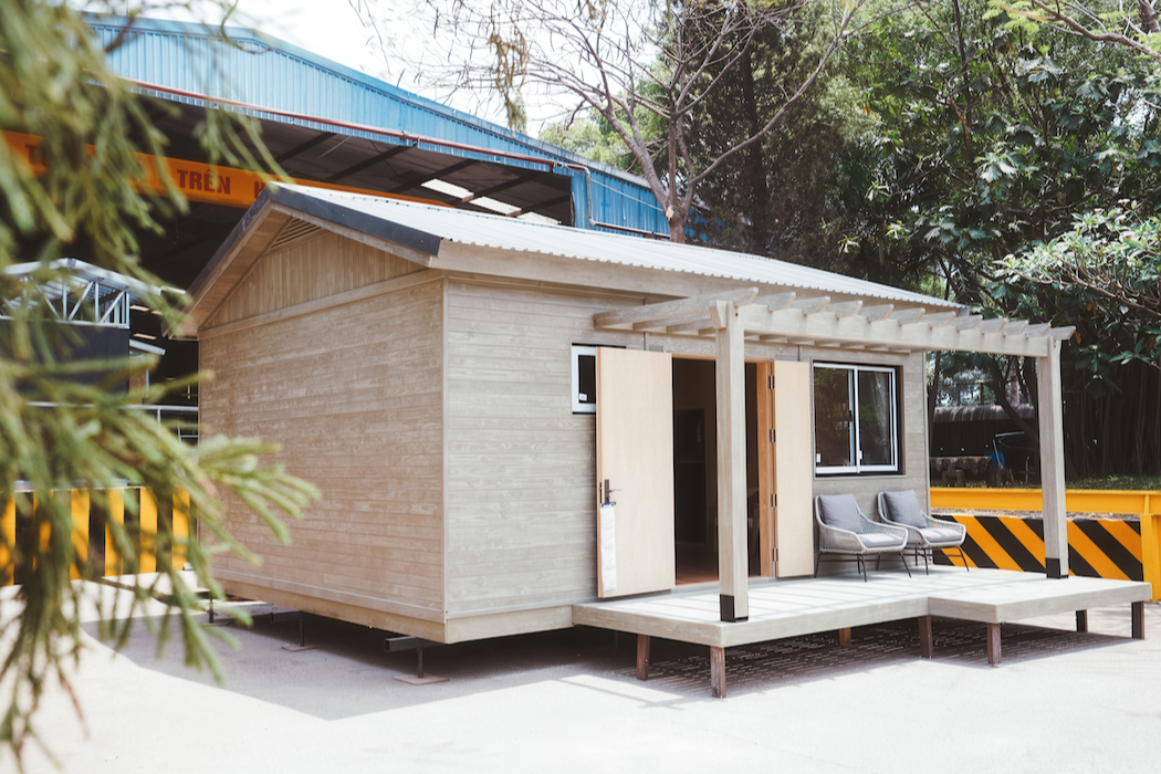 A small wooden house with a porch featuring two chairs, situated outdoors with trees in the background.