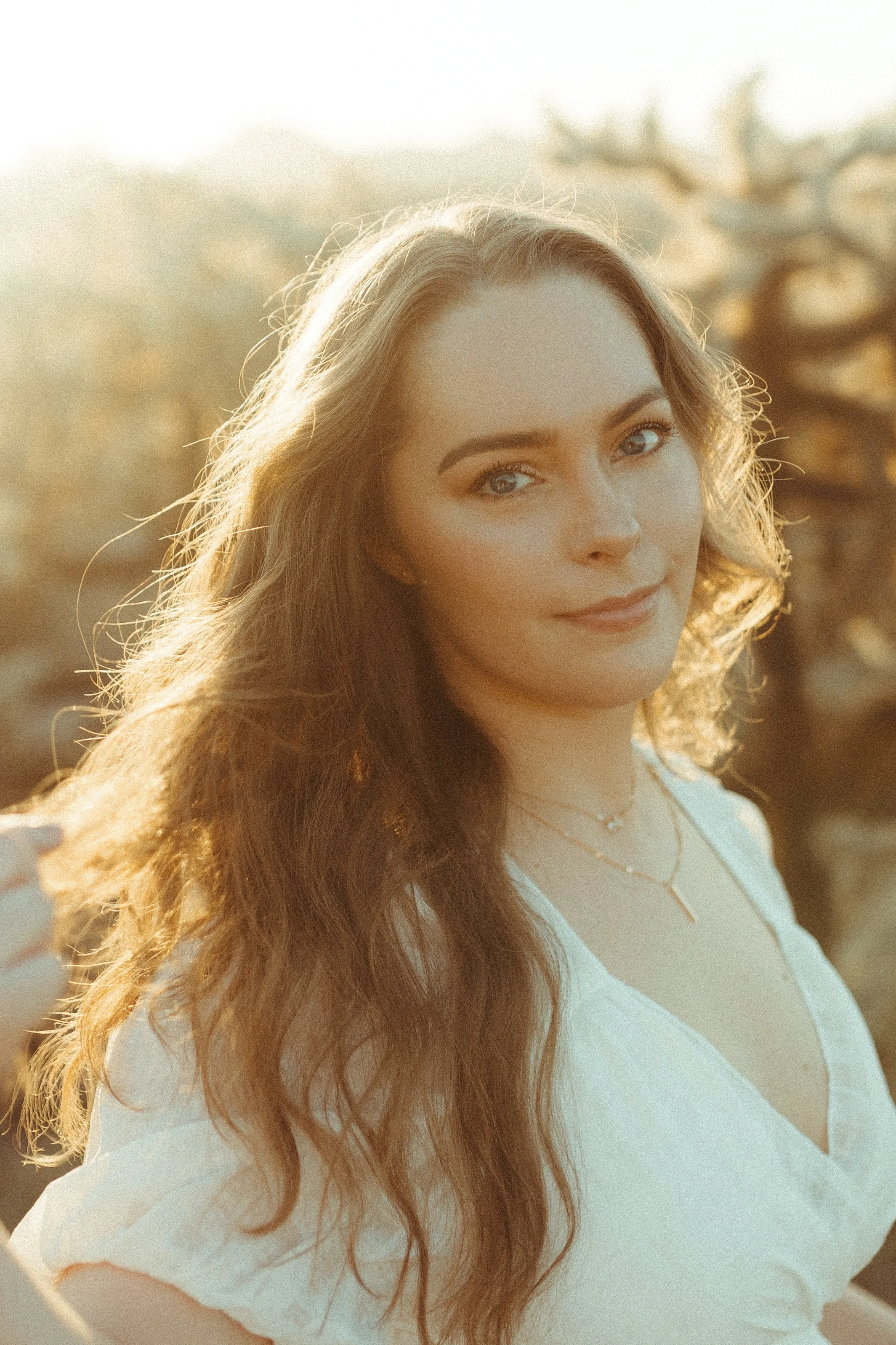 A young woman with wavy brown hair and light makeup, smiling gently, outdoors in sunlight, wearing a white top and layered necklaces.