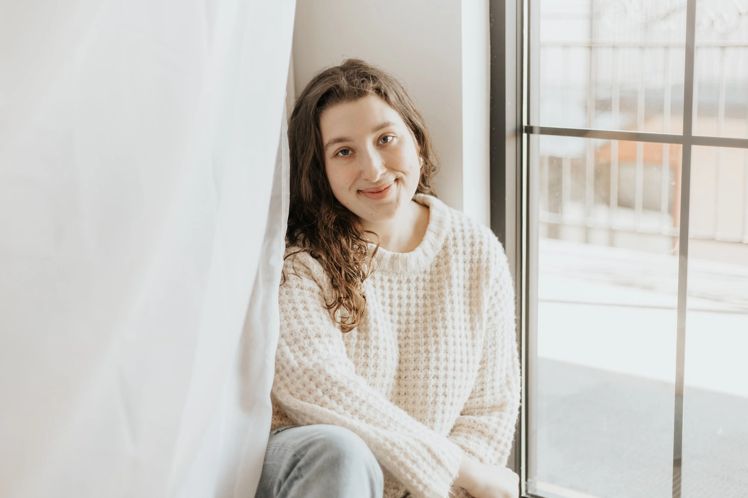 Young woman with curly brown hair sitting by a window, smiling at the camera, wearing a light-colored knitted sweater.