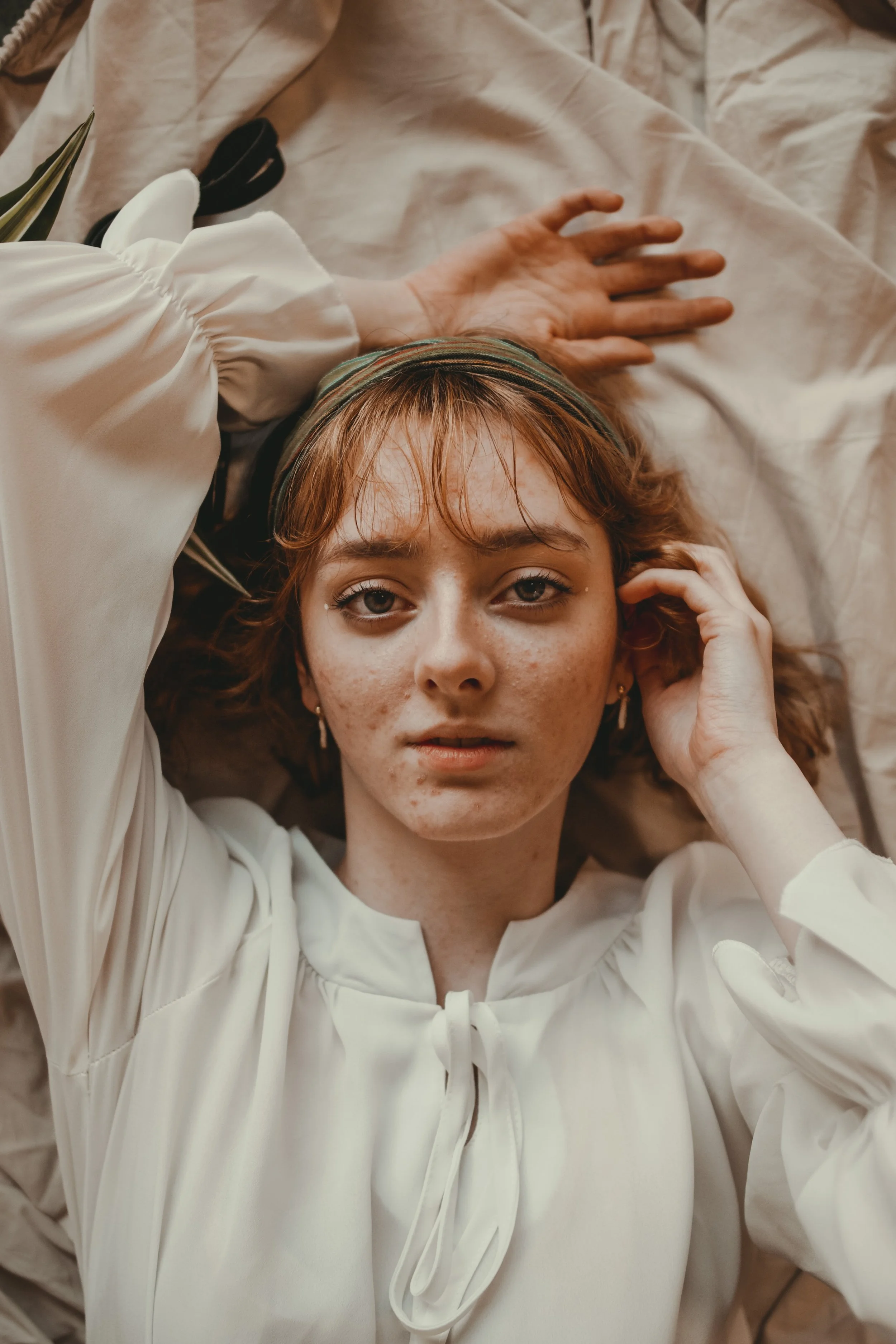 A young woman with freckles and red hair lying on a beige bed, looking into the camera, wearing a white blouse and a striped headband.