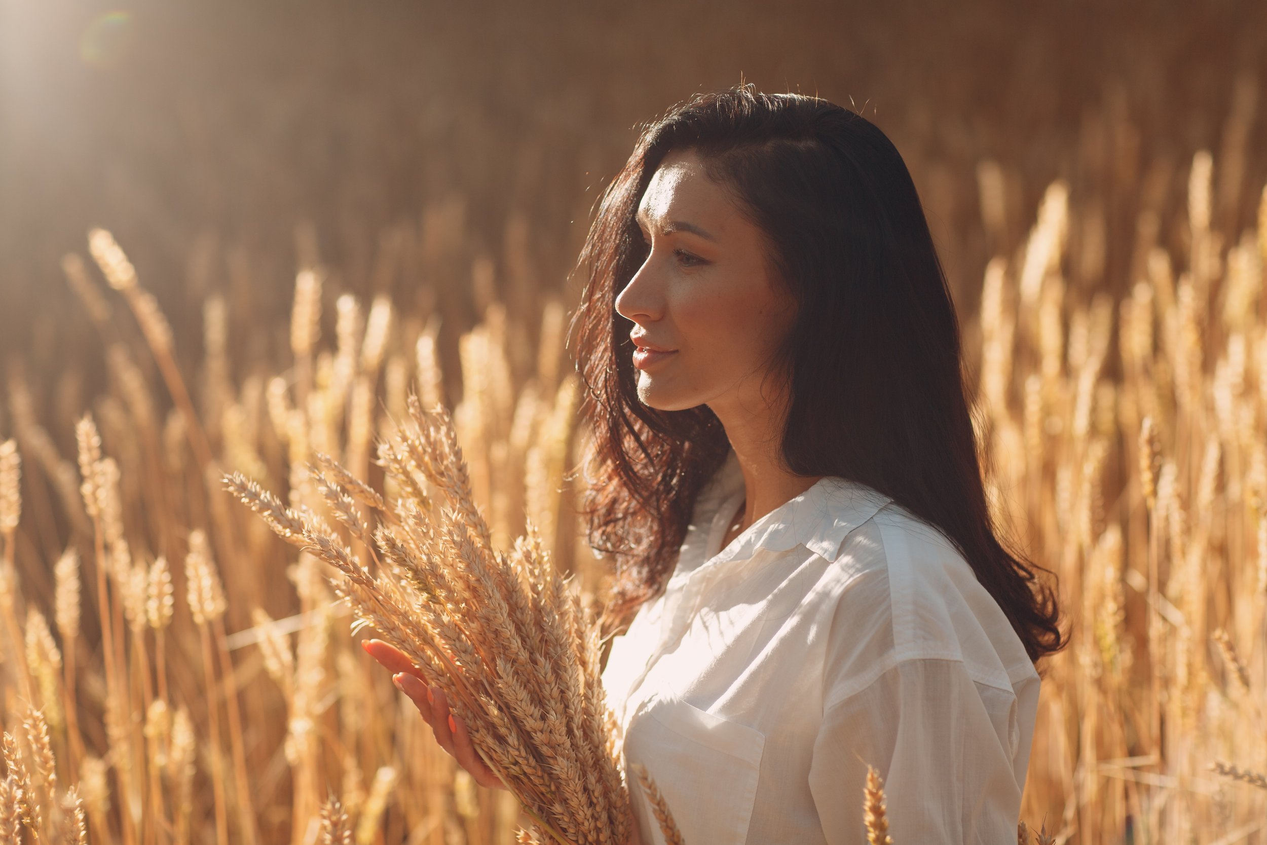 A woman with long dark hair holding a bundle of wheat in a golden wheat field at sunset.
