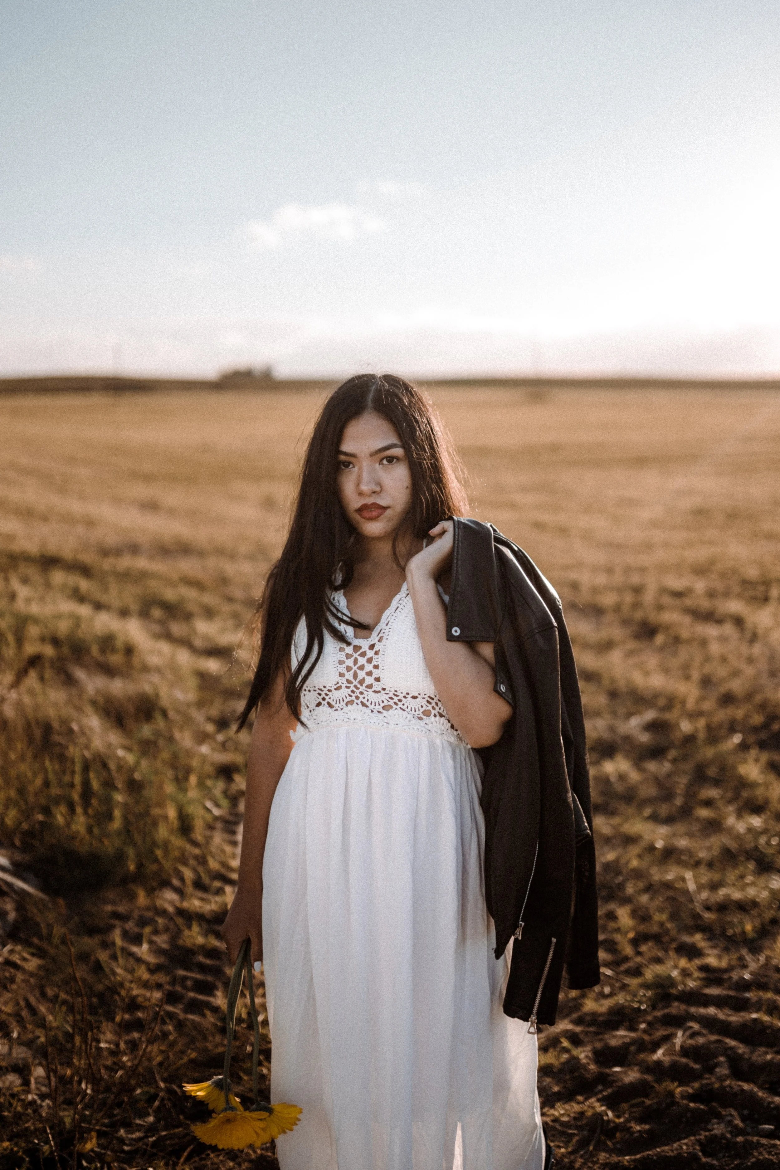 A woman in a white dress holding a sunflower in one hand and carrying a black leather jacket over her shoulder stands in a field during sunset, with an open sky in the background.