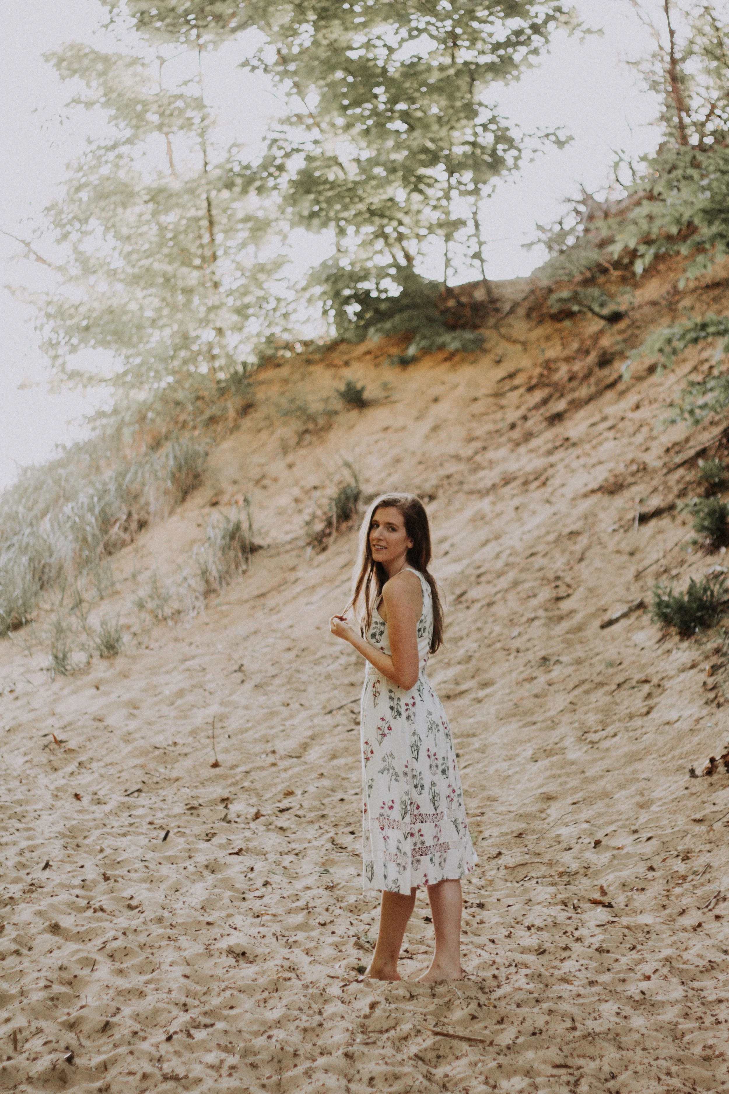 A woman in a white floral dress walking barefoot on a sandy slope with trees in the background.
