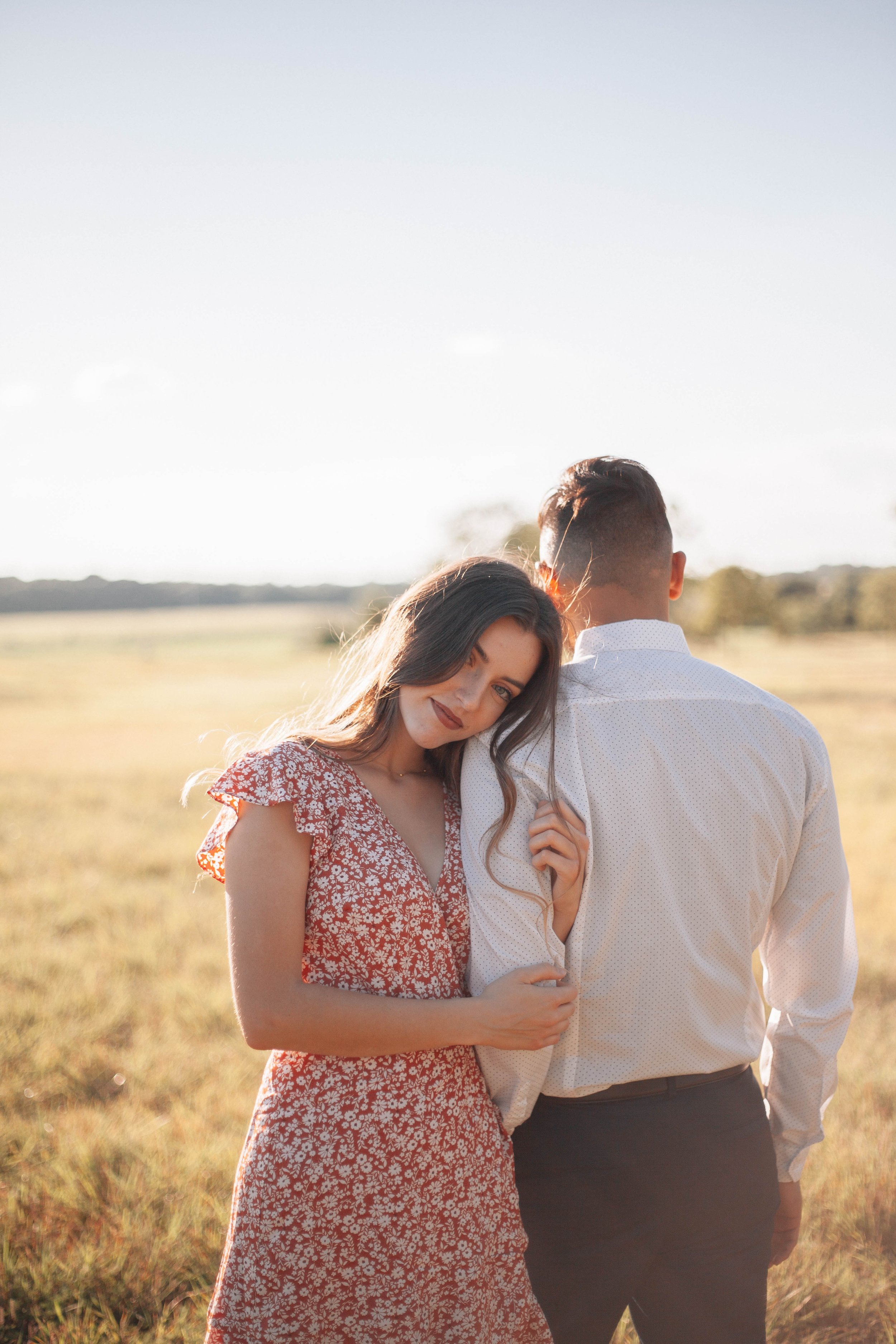 A young woman with wavy brown hair and a red floral dress embraces a man in a white shirt in a sunny, open field.