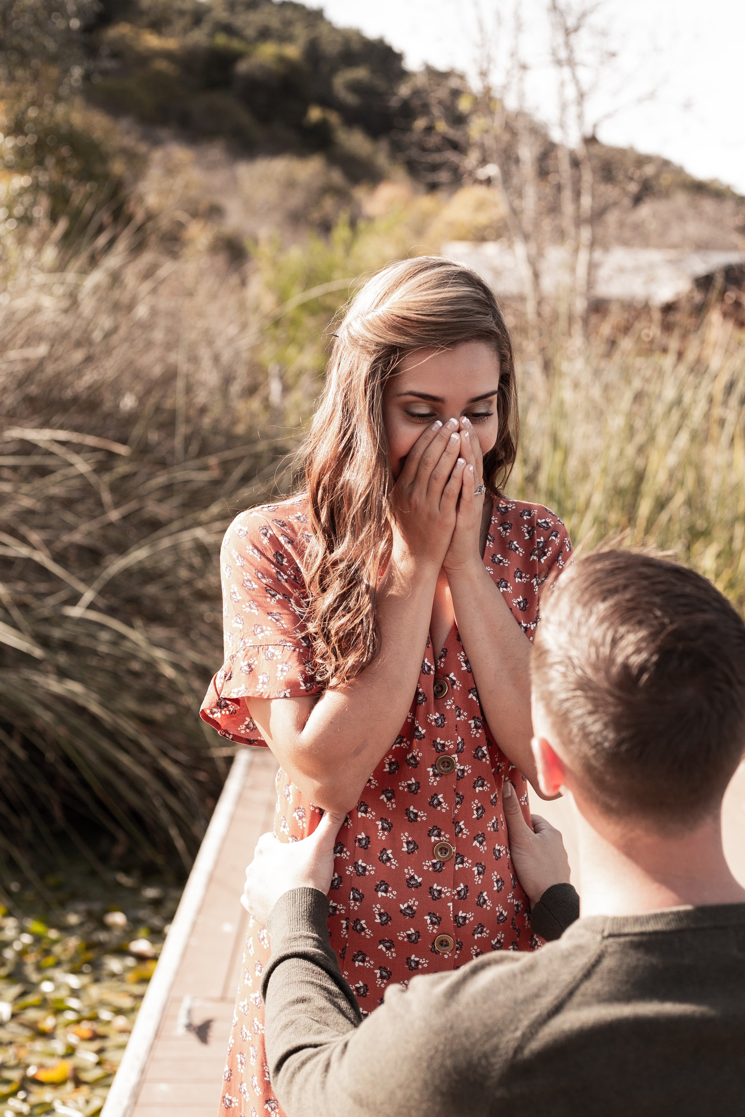A man proposing to a woman on a wooden dock outdoors, with the woman covering her mouth in surprise and joy.