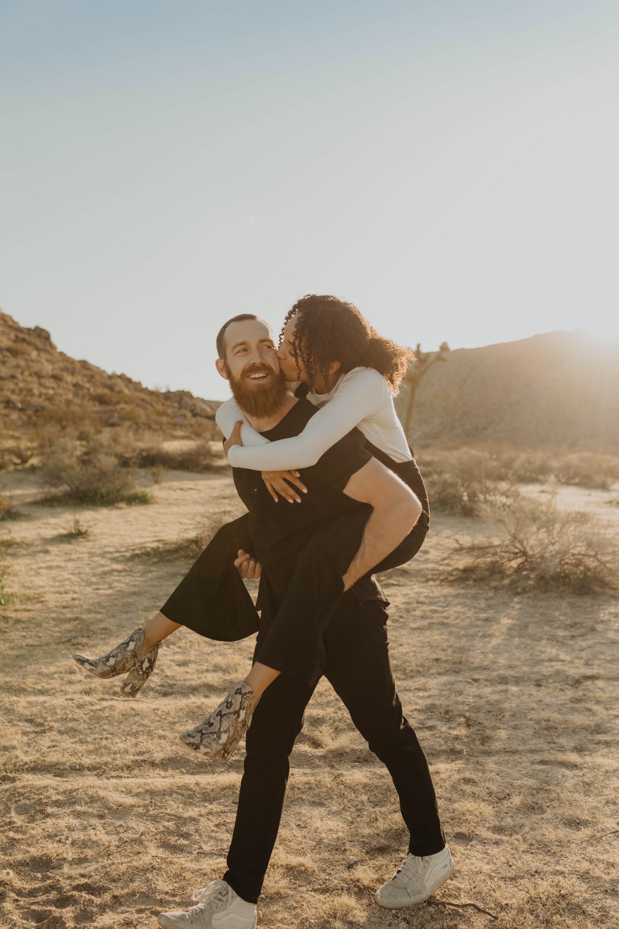 A man carrying a woman on his back in a desert landscape during sunset, both smiling and embracing affectionately.