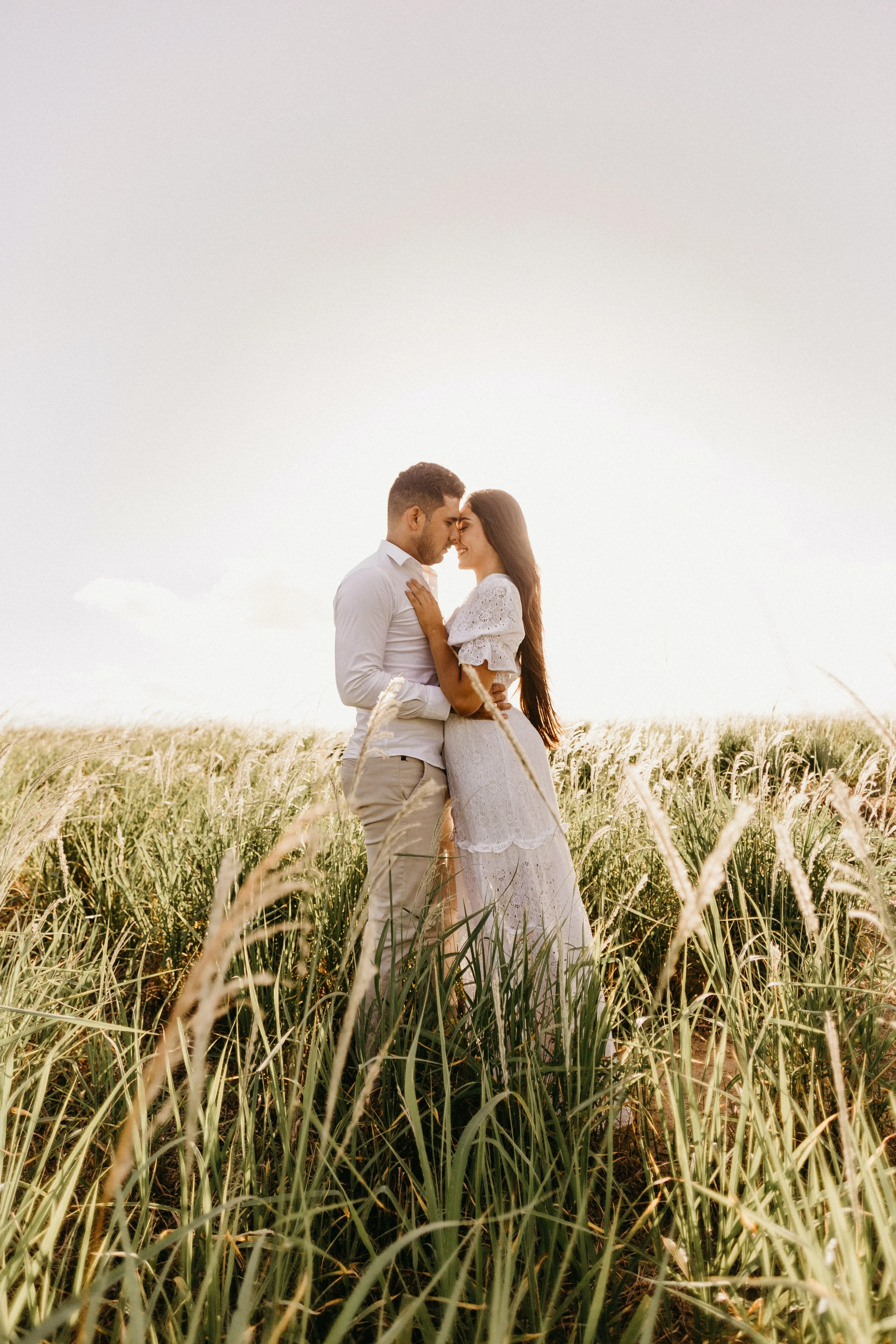 A couple standing close together in a field of tall grass, holding each other and touching foreheads during sunset.
