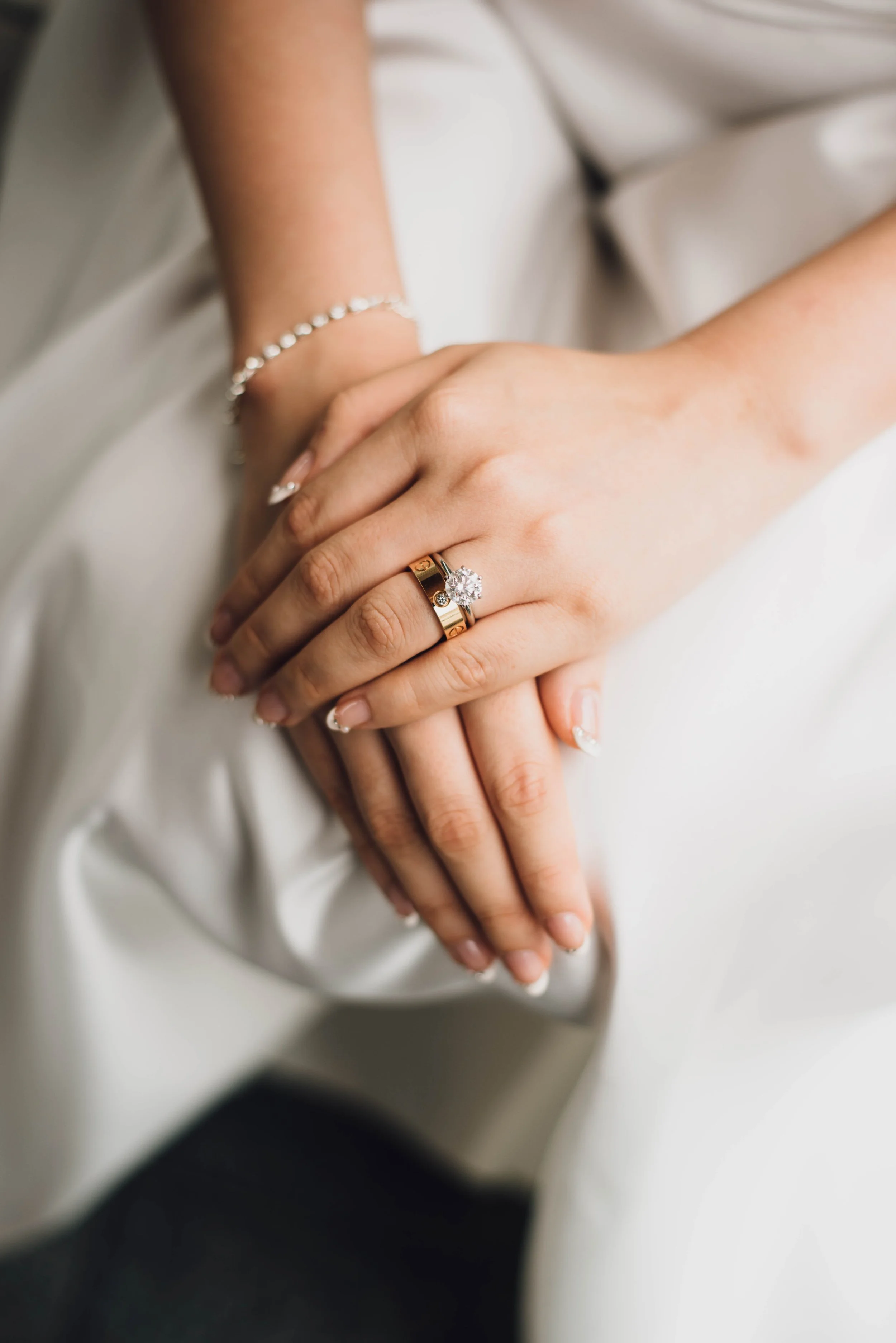 Close-up of a woman’s hands showing her wedding ring with a large diamond solitaire, resting on her lap, with another hand holding hers, wearing a bracelet and neatly manicured nails.