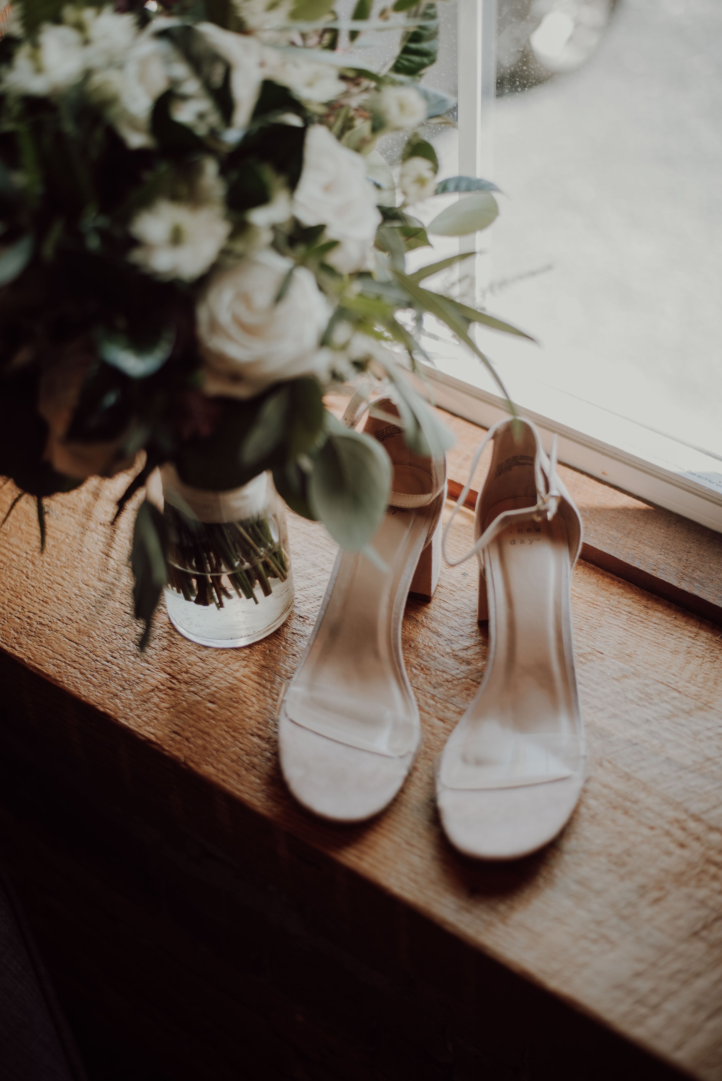A pair of white high-heeled shoes on a wooden surface next to a vase with white and green flowers, near a window with natural daylight.