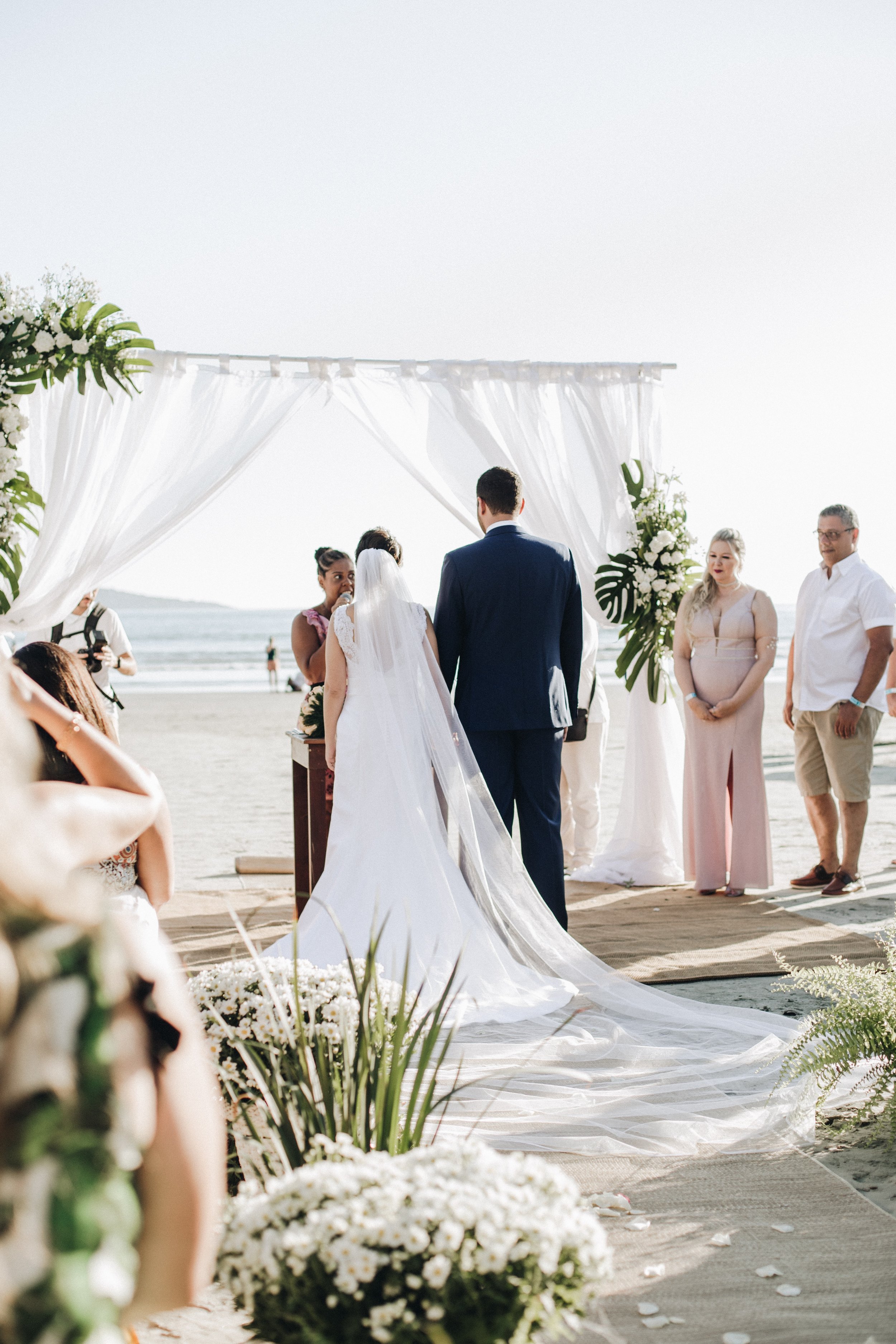 A beach wedding ceremony with a bride and groom standing under a floral arch with white drapes, on sandy ground near the ocean.