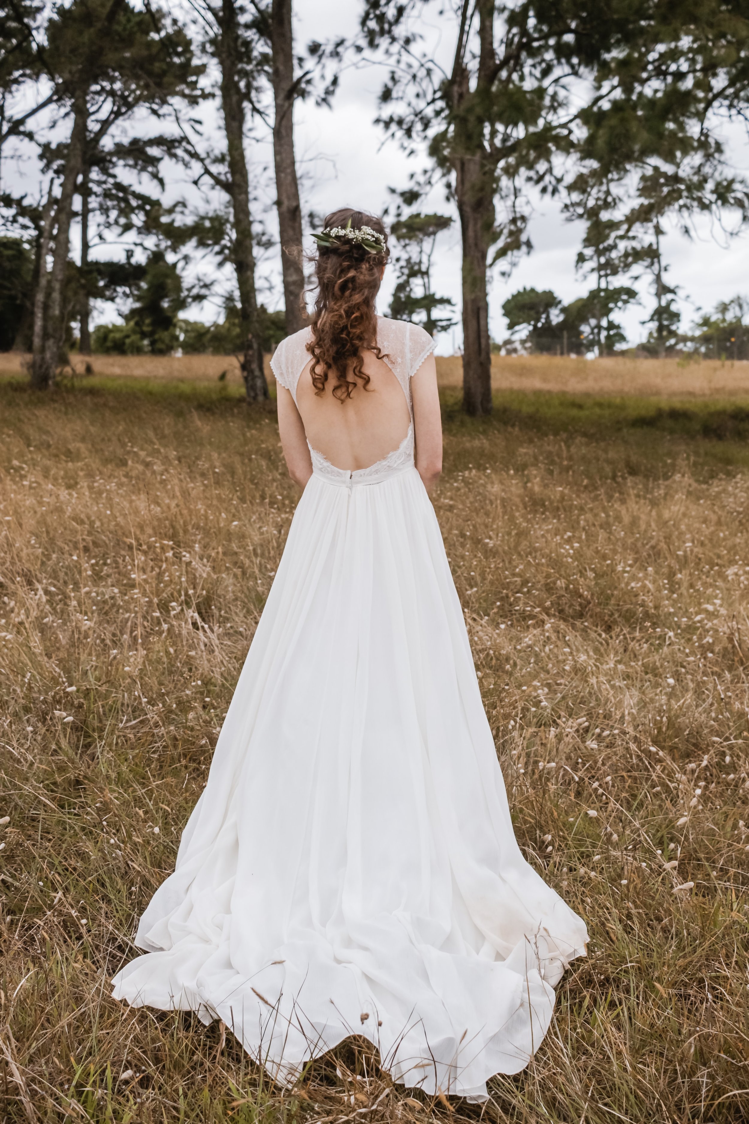 Back view of a woman in a white wedding dress with open back standing in a grassy field amidst trees.