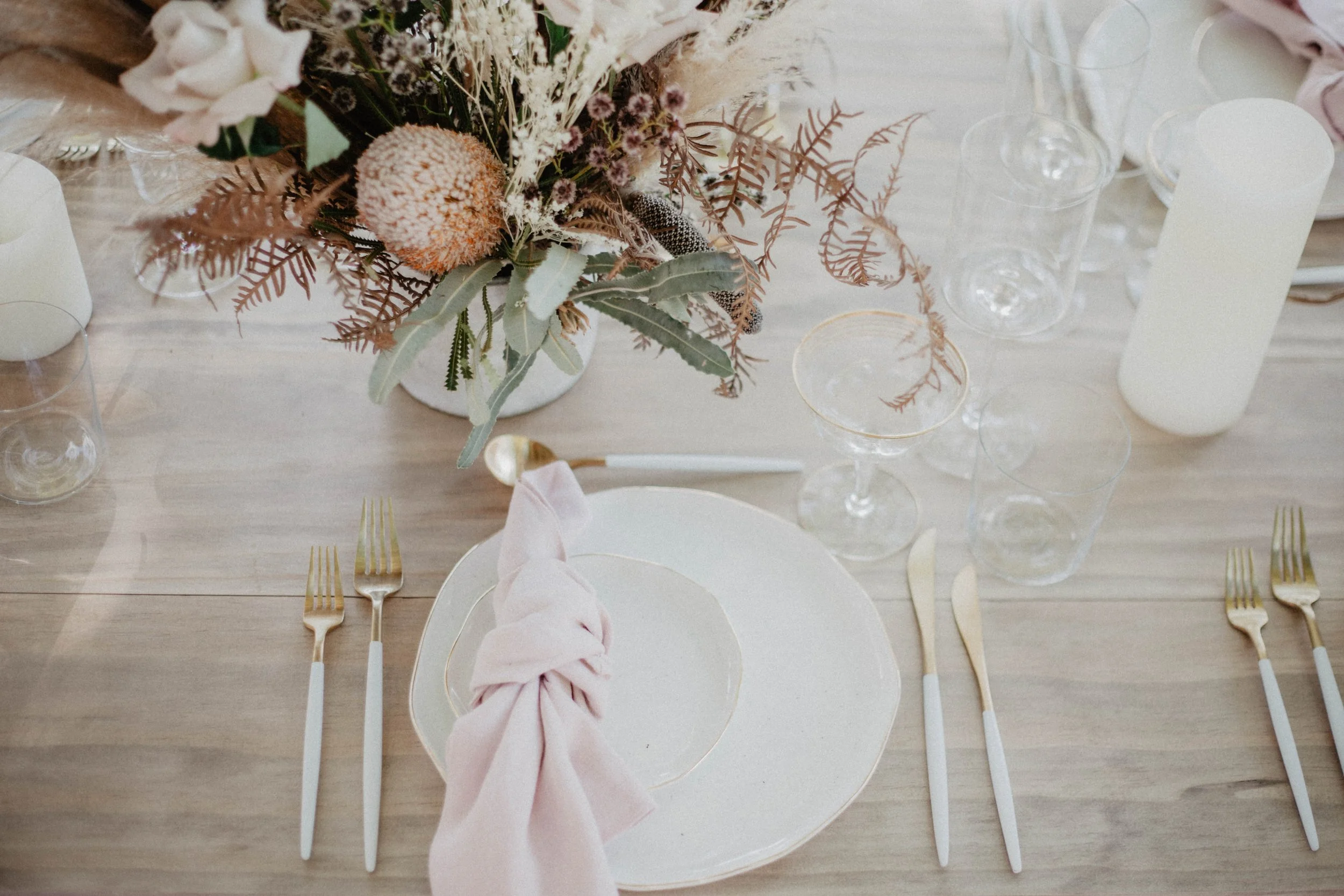 Elegant table setting with a floral centerpiece, gold and white flatware, pink napkin tied on a white plate, and multiple clear glassware on a light wood table.