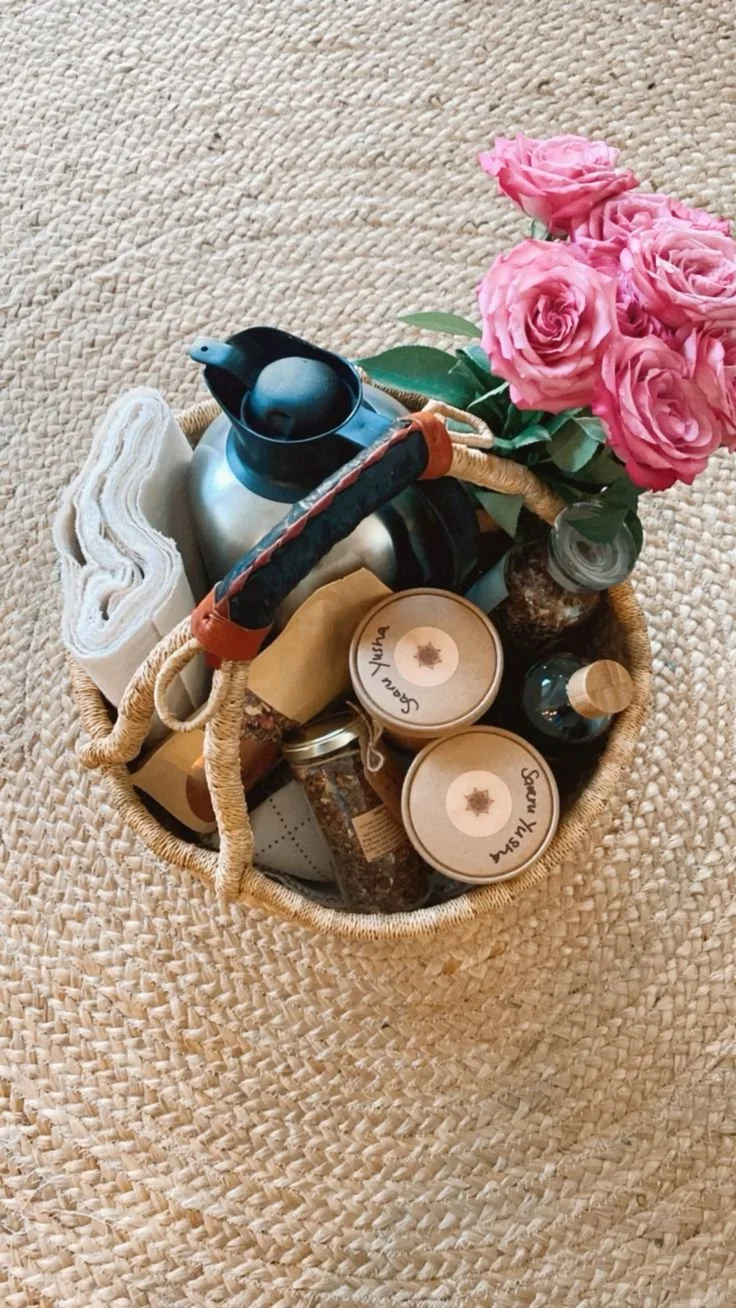 A woven straw basket containing a pink rose bouquet, a white towel, a stainless steel thermal carafe, a black and orange tennis racket, small jars labeled 'Scvan Yusha,' a glass jar, and a small container, placed on a textured beige rug.