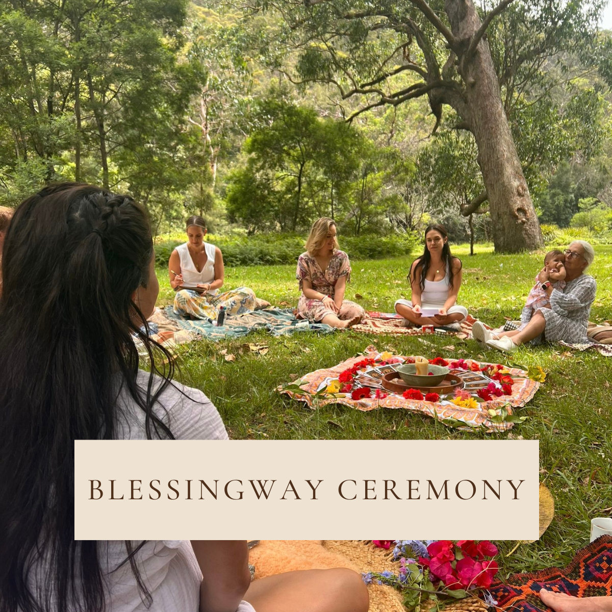 A group of women and a young girl participating in a blessingway ceremony outdoors in a lush, green park with large trees. They are sitting on colorful blankets in a circle, with a decorated central altar featuring flowers, candles, and religious or spiritual items, celebrating a special occasion.