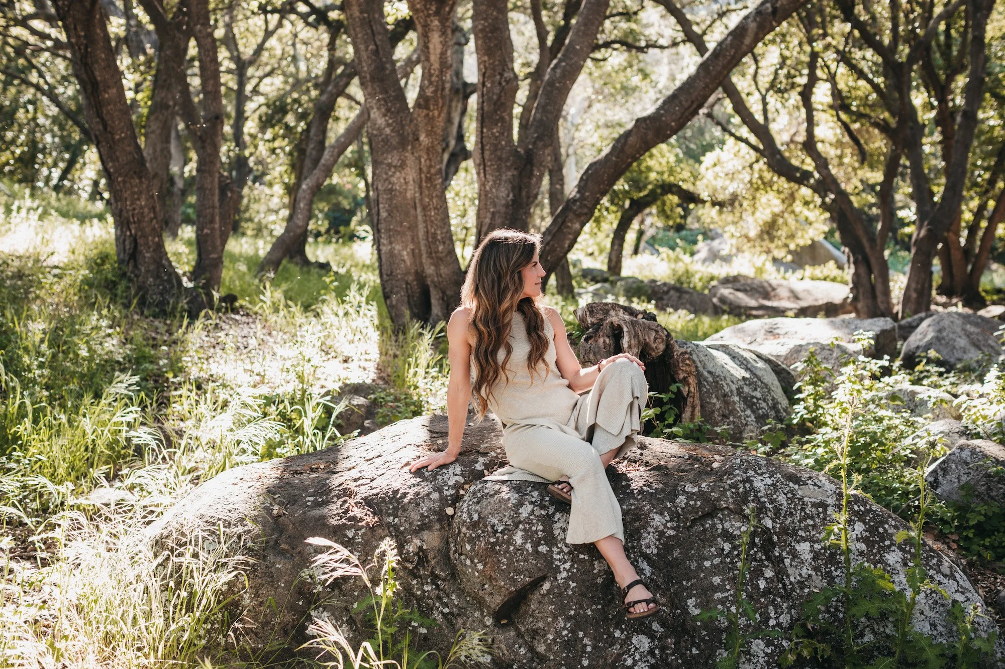 A woman with long wavy hair in light-colored clothing sitting on a large rock in a forested area with tall trees and greenery, bathed in sunlight.