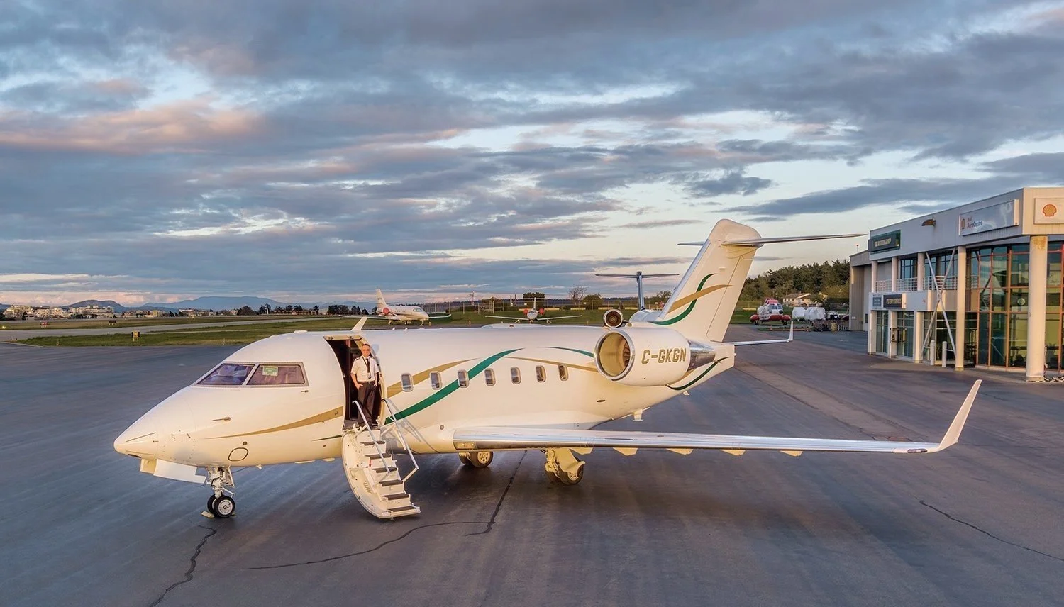 Small private jet with open door and crew member standing near stairs on tarmac, airport building in the background, blue sky with clouds.